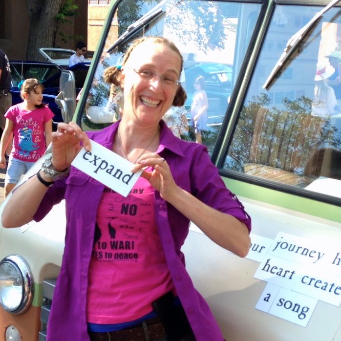 Jennifer Rayman holding the sign "Expand" she has what appear to be pigtails in her hair and is wearing a fuscia shirt over a short sleeved pink t-shirt with black lettering: with "Women say No to War"  and No in various languages