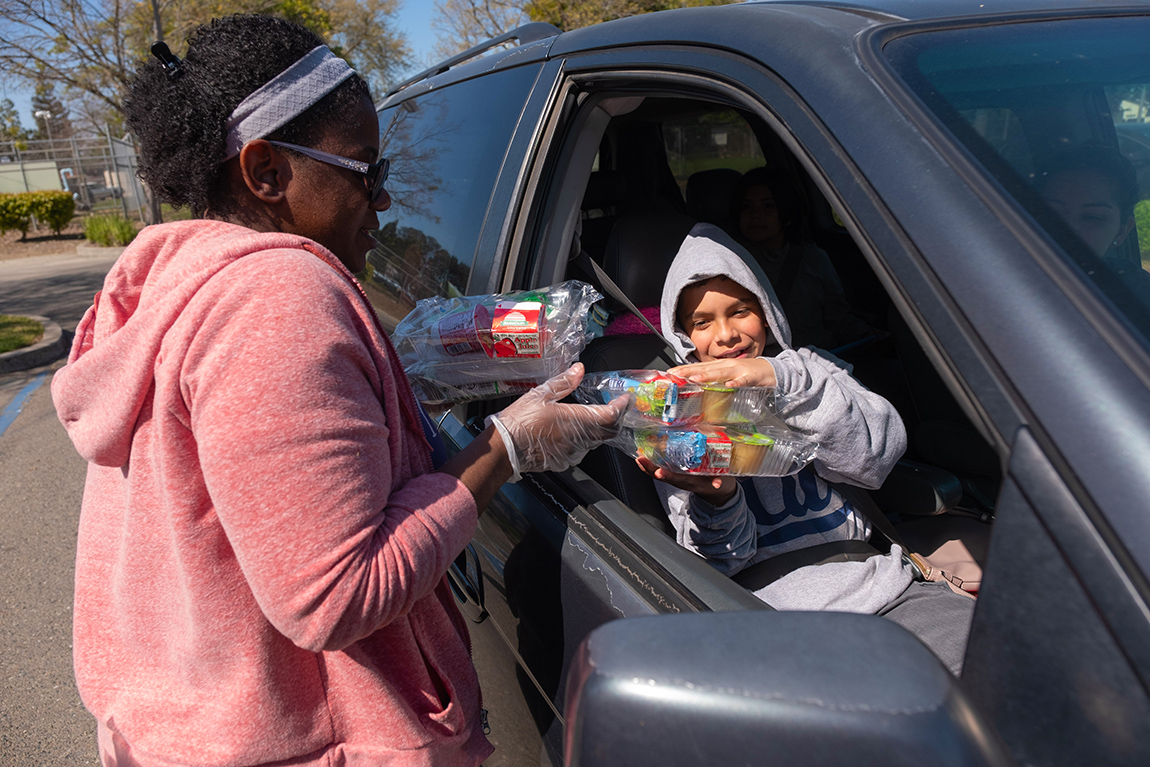 A woman hands a boxed lunch to a student through an open car window.