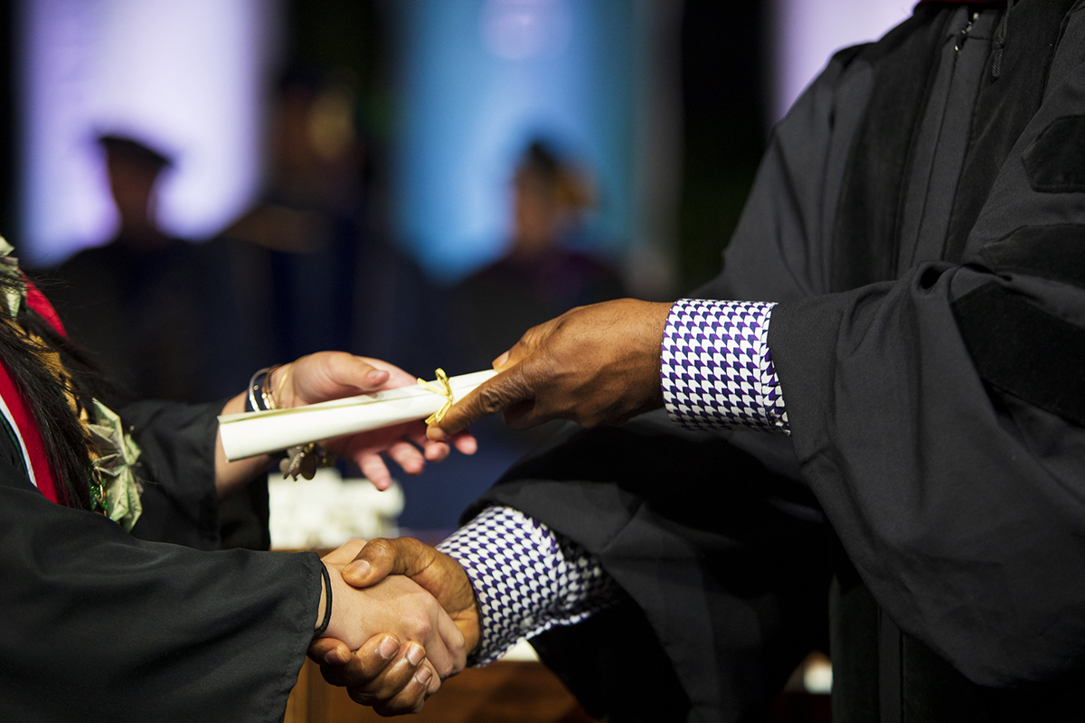 A person in academic robe handing a college degree to another person in a graduation gown.