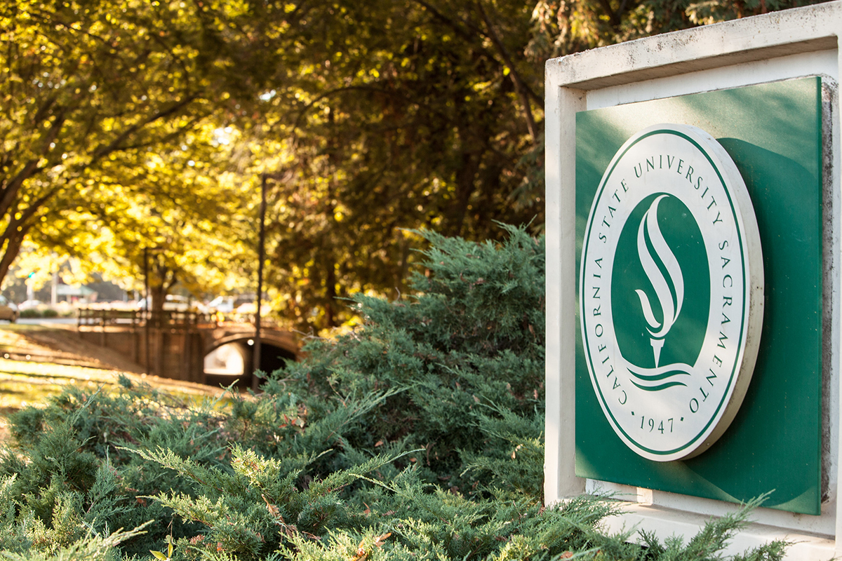 A photo of the CSUS sign at the front of campus, with a walkway and trees in the background