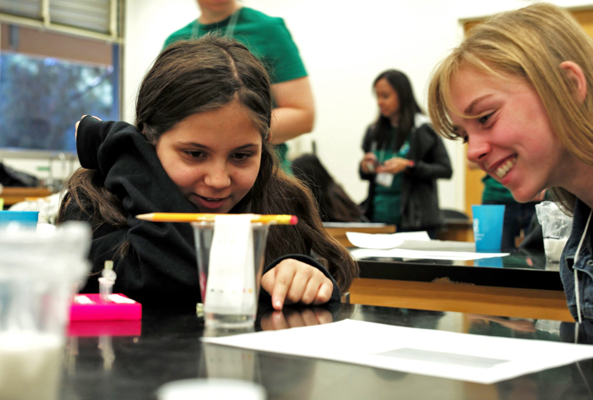A girl in a science lab looking at a pencil being suspended over a glass cup