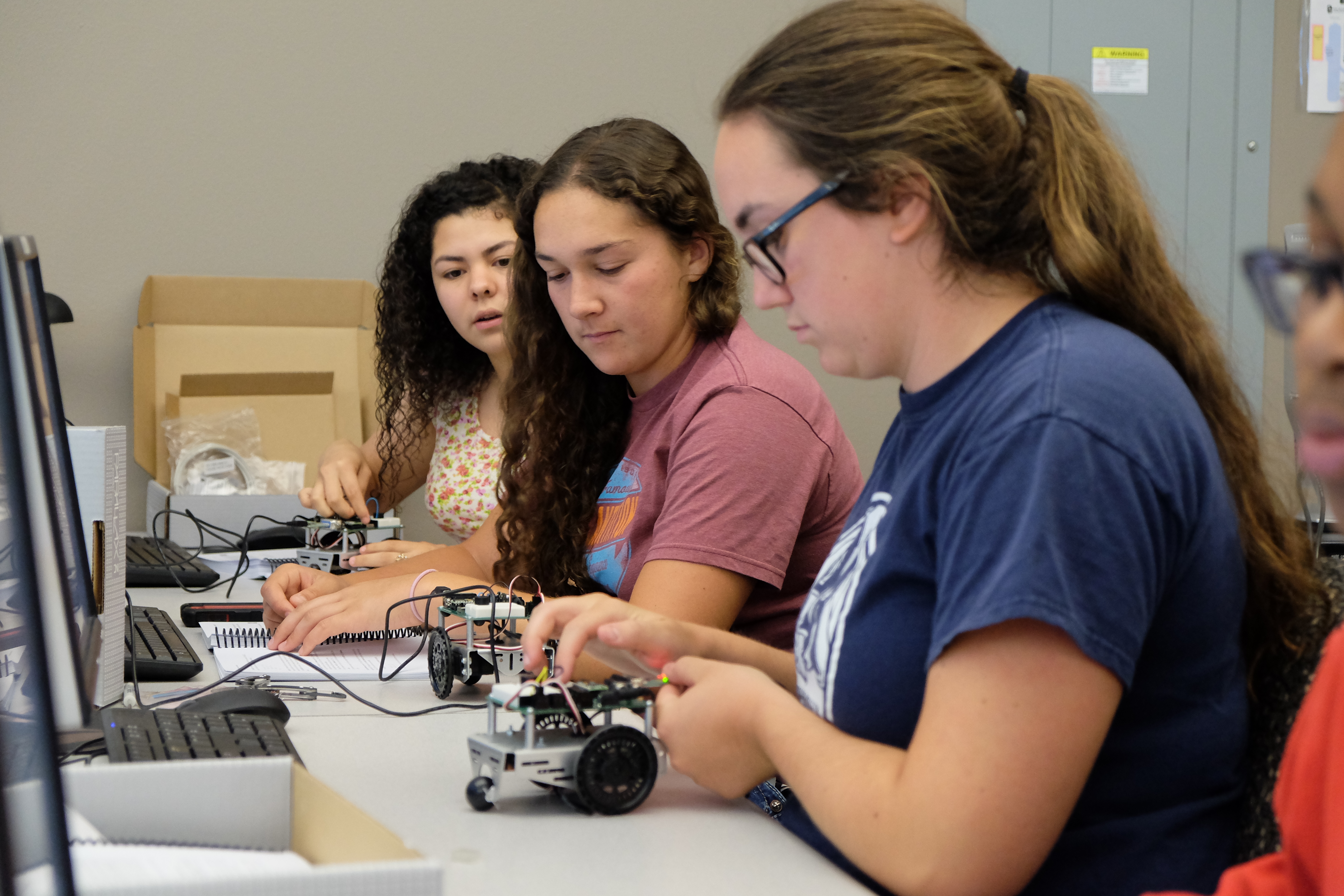 A photo of three high school students at a desk working on small robots.