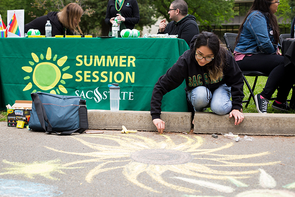 A photo of a student drawing on the pavement with chalk, in front of a table with a green banner reading 'Summer Session'