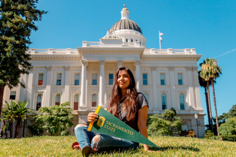 Student sitting on the grass in front of the California State Capital building holding a green Sac State pennant.