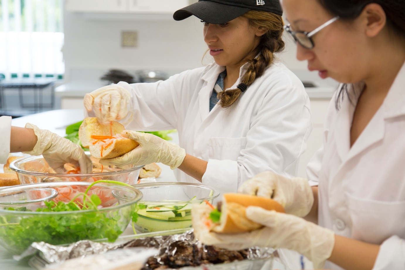 A female student in a white lab coat with a braided side ponytail and a black baseball cap stands behind another student in a white lab coat wearing glasses. Both students are prepare sandwiches. In the background is a blurred kitchen setting.