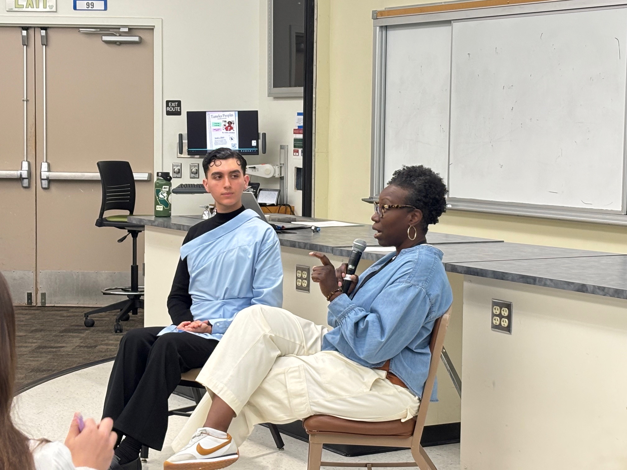 Two people sitting in front of a large classroom. One person is holding the mic and speaking into it and the other person is listening.