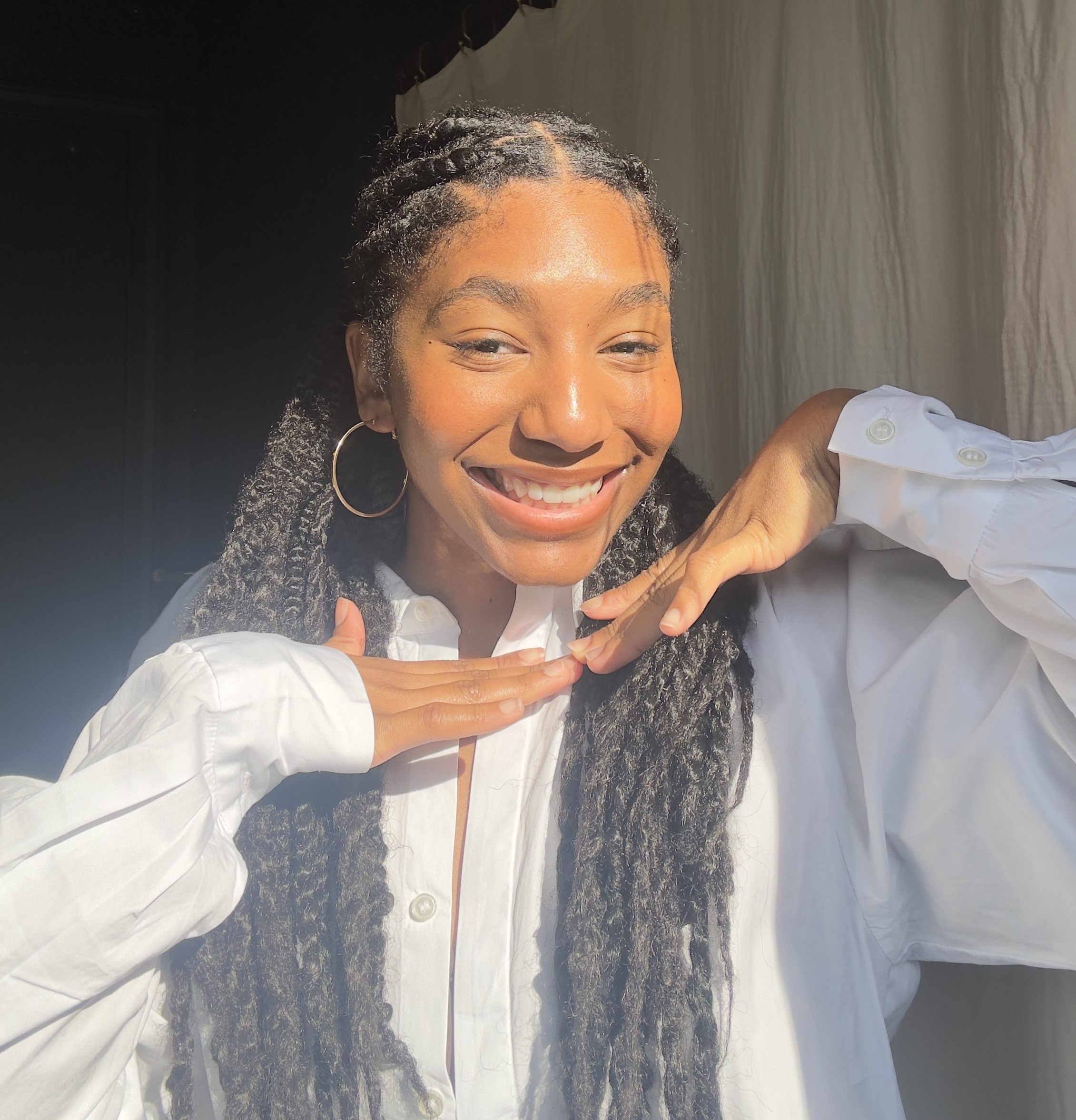 Light skinned Black female with long twists and gold hoop earrings wearing a white button down blouse smiles while holding hands together under her chin to frame her face