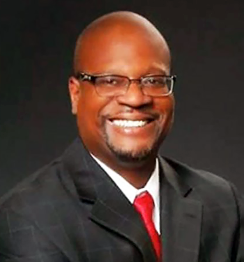 Skip Bishop smiles, wearing black eyeglasses, a black suit with a white shirt and red tie. 