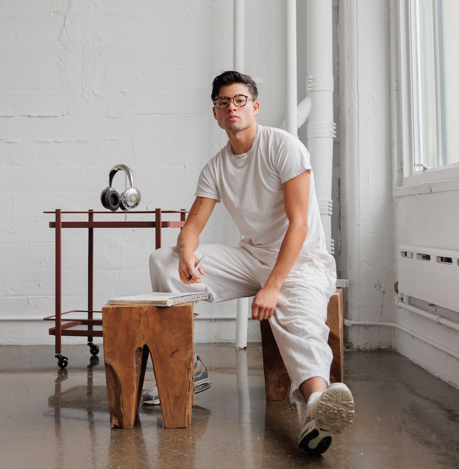 Chella Man wearing glasses and holding a marker in his right hand in a room with white walls and brown floor in white shirt and pants sitting on a brown wooden stool with one leg in front and the other bent. He sits behind another wooden stool that has a notebook on top and to the right of a pair of silver headphones from a brown rolling cart.