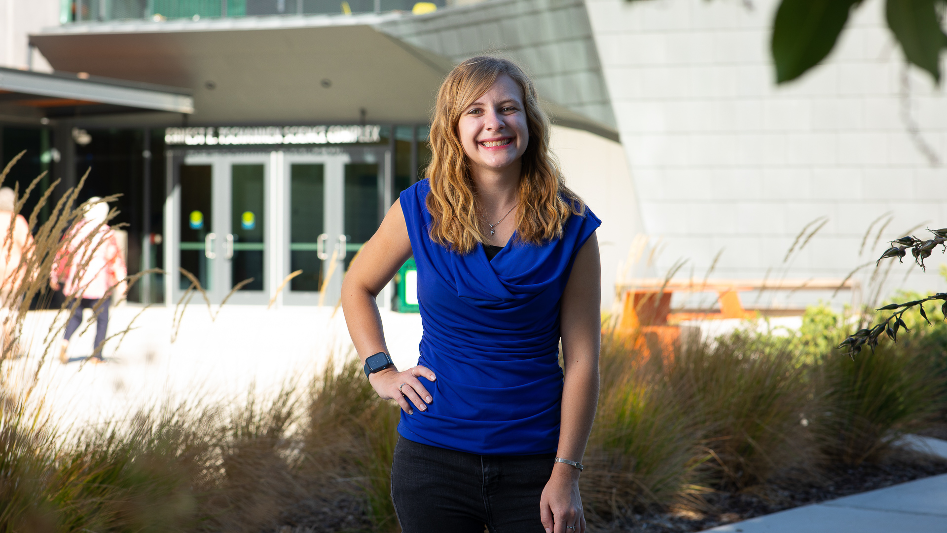 Sac State alumna Elizabeth Gabler, who worked on the Double Asteroid Redirection Test (DART) project at Sacramento-based aerospace and defense firm Aerojet Rocketdyne, stands in front of the Ernest E. Tschannen Science Complex with a big smile while wearing a blue shirt and black pants.