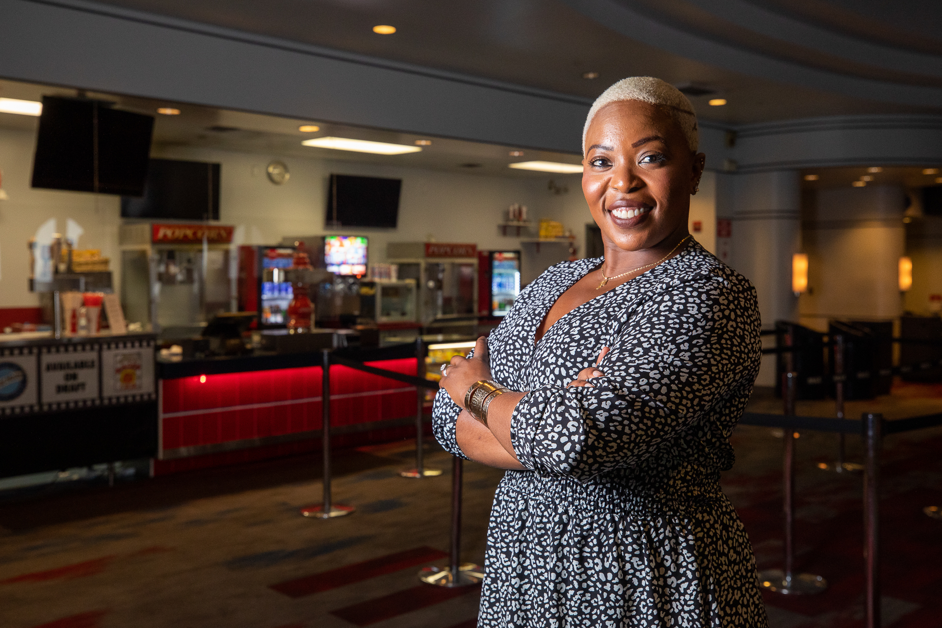 Melissa Muganzo Murphy, standing with arms crossed, looking at the camera, in the lobby of a movie theater