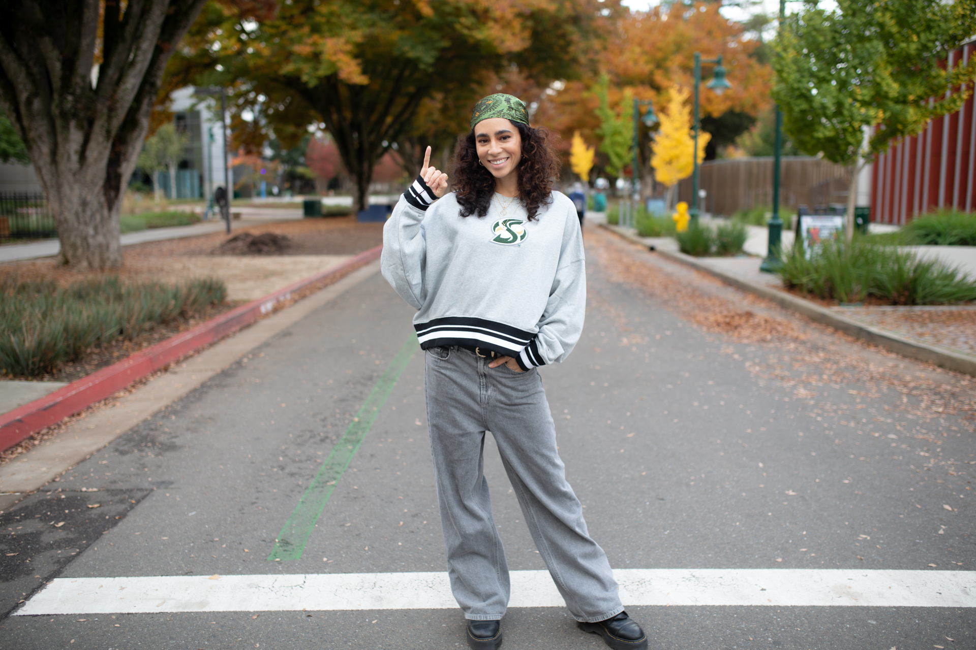 A woman in Sac State sweatshirt poses on campus with her pinky raised in Stingers Up sign.