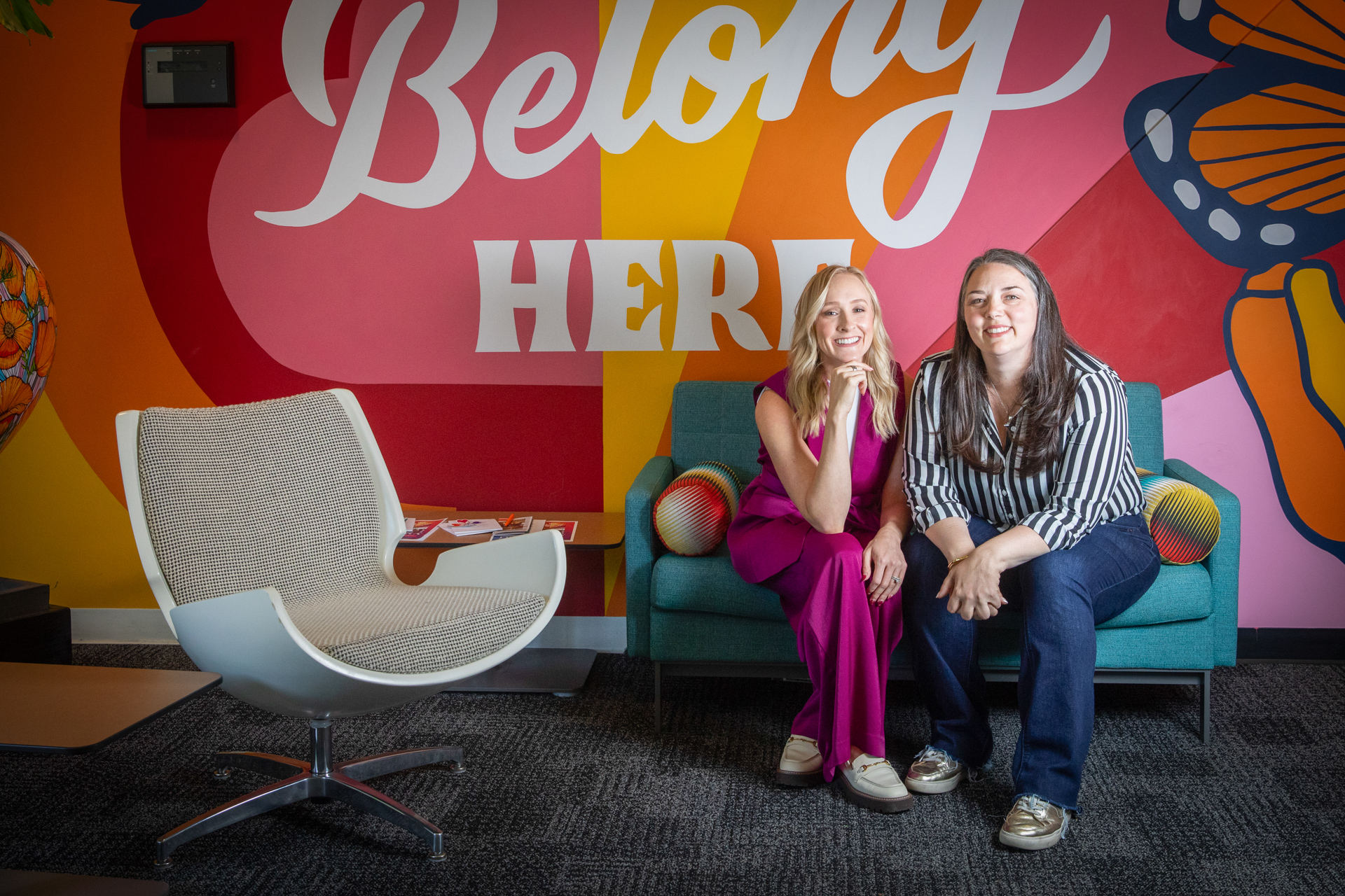 Anne Descalzo and Rachel Zillner sitting in front of a colorful wall reading "Belong Here."