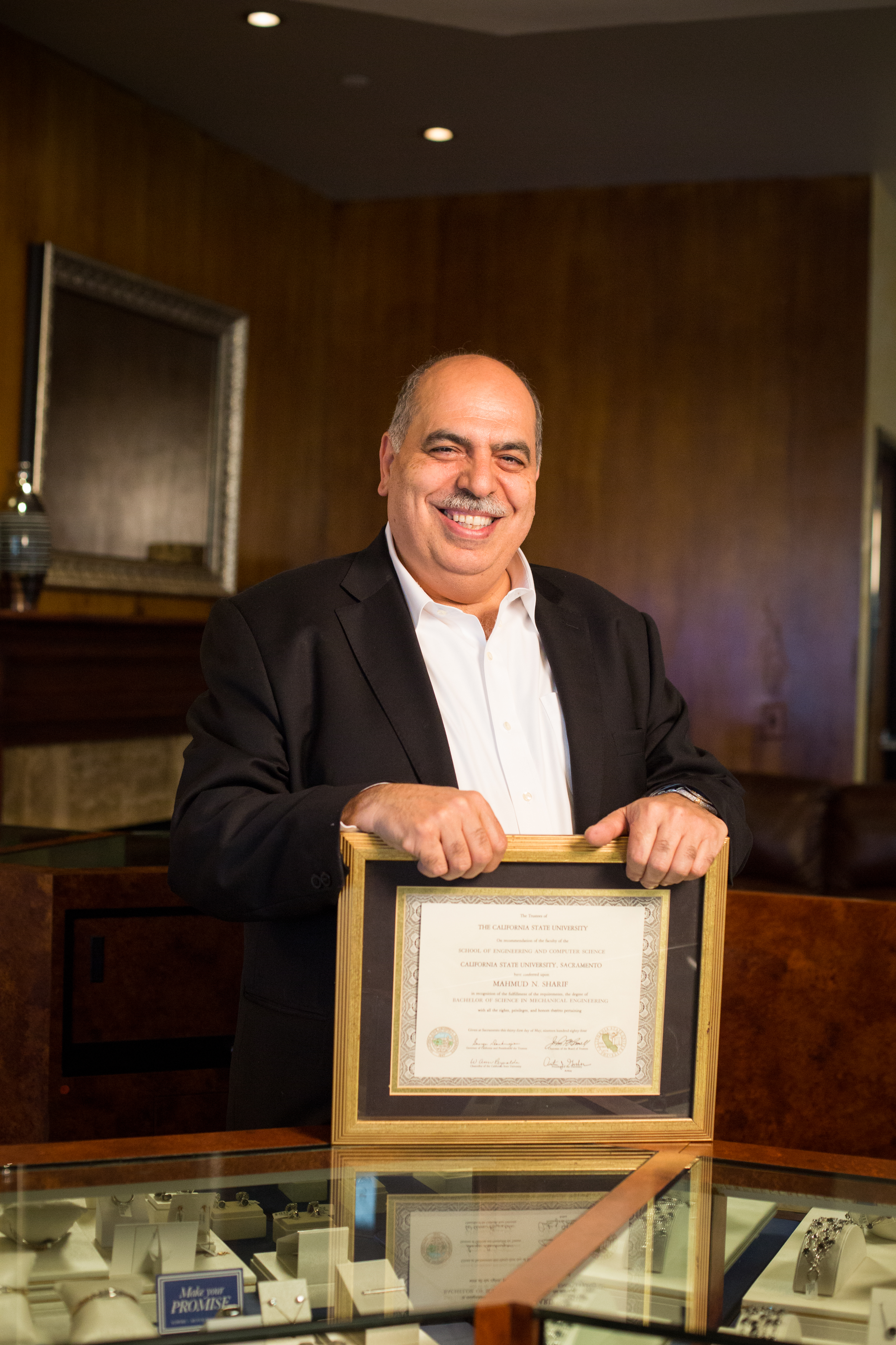 Mahmud Sharif posing for a photo with his Sac State diploma, standing behind a glass display case.