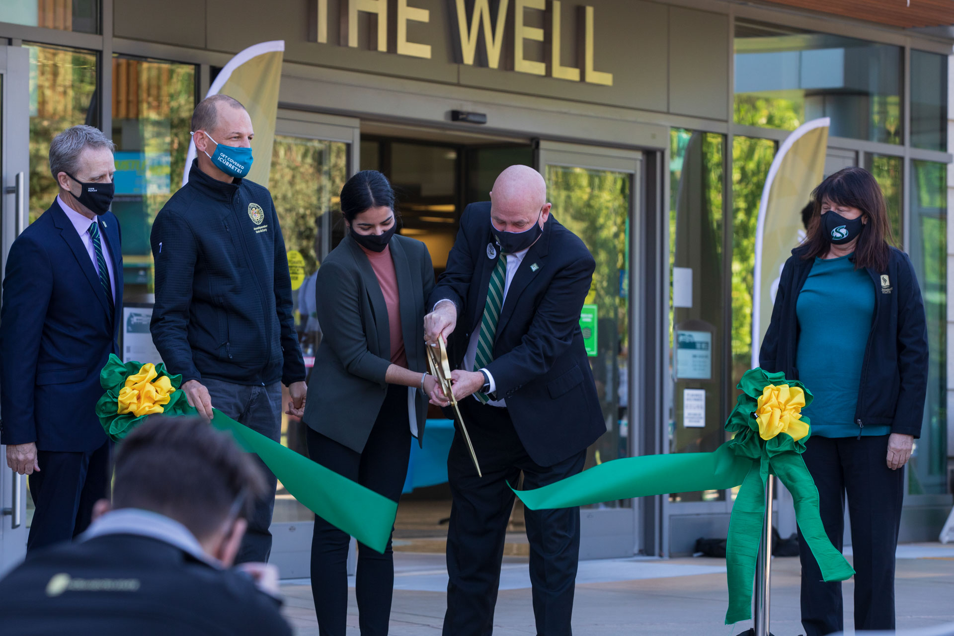 Two individuals cut a green ribbon with a large, gold pair of scissors, as three others look on, in front of The WELL