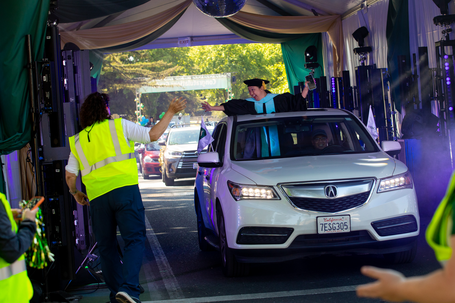 A Sac State graduate in a cap and gown stands up in the passenger seat of a car as it passes through a tent filled with lights and sound equipment.. 
