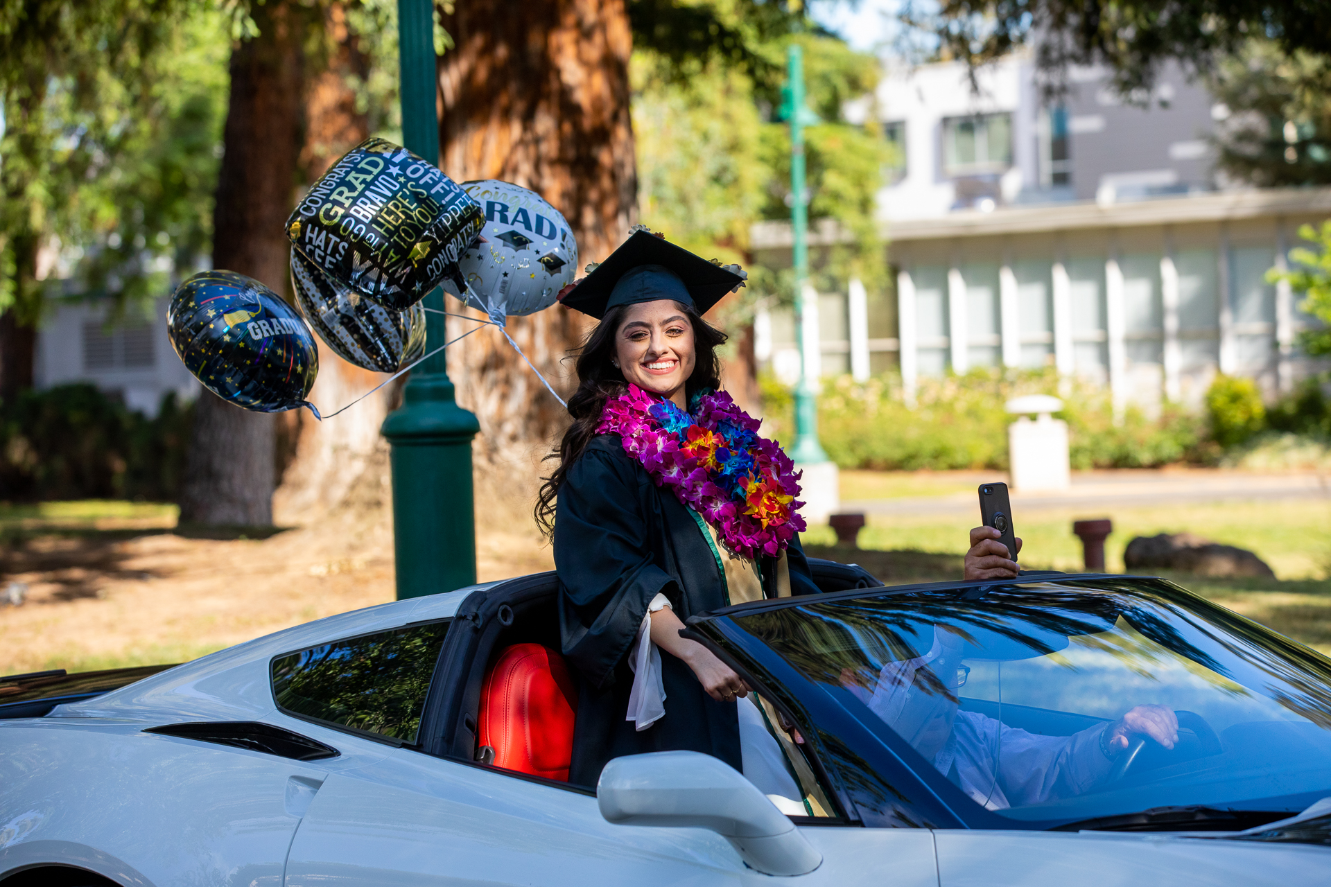 A car decorated with a flag reading "Congrats Grad" drives through campus, as people cheer from the sidewalk.