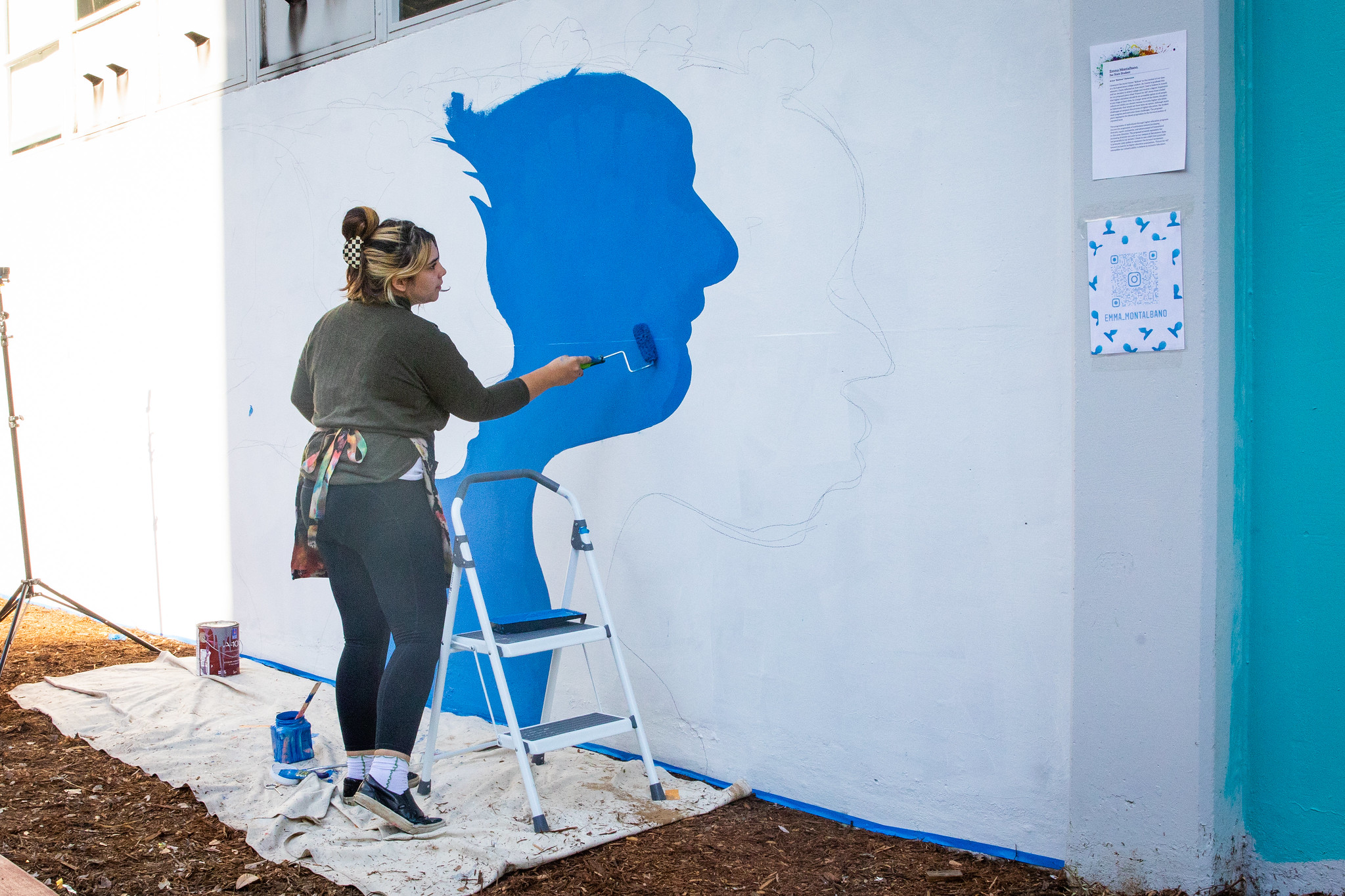 Emma Montalbano, standing outdoors, painting a mural depicting a person in side profile, colored blue on a white background