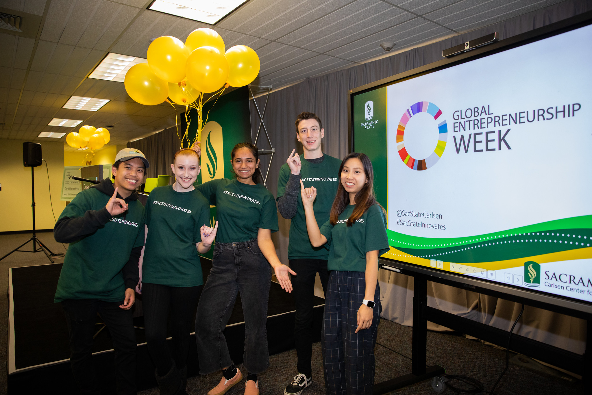 Five students in green shirts reading "#SacStateInnovates," standing next to a digital display reading "Global Entrepreneurship Week."