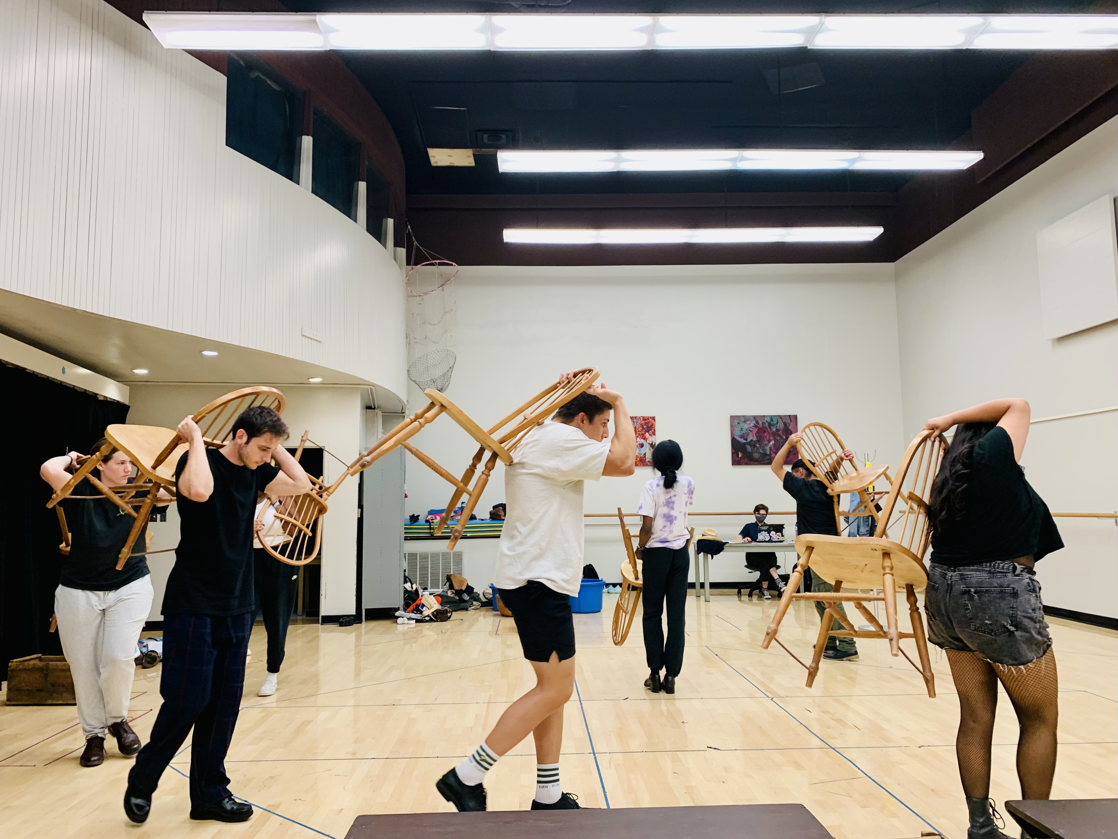 Actors in a rehearsal space, walking in a circle carrying wooden chairs.