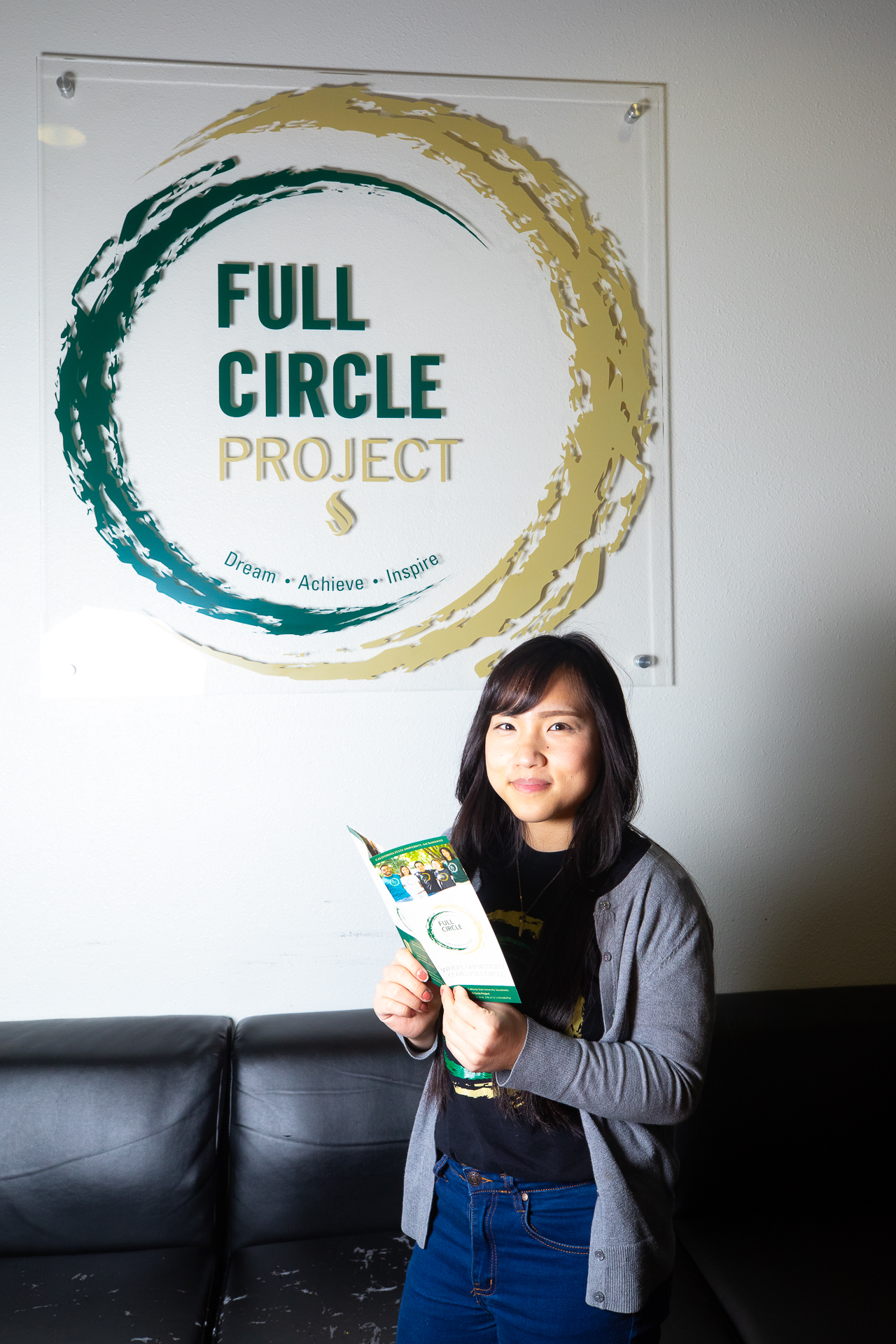 Sacramento State student Hnou Lee, standing indoors, in front of a Full Circle Project logo on the wall, holding a Full Circle Project brochure