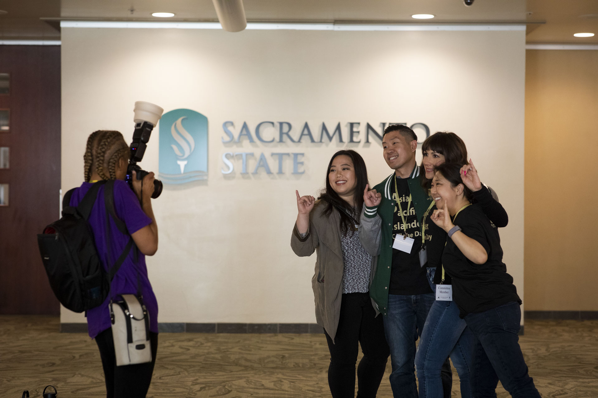 A photographer taking a picture of four students, standing, indoors, in front of a wall with the Sacramento State logo