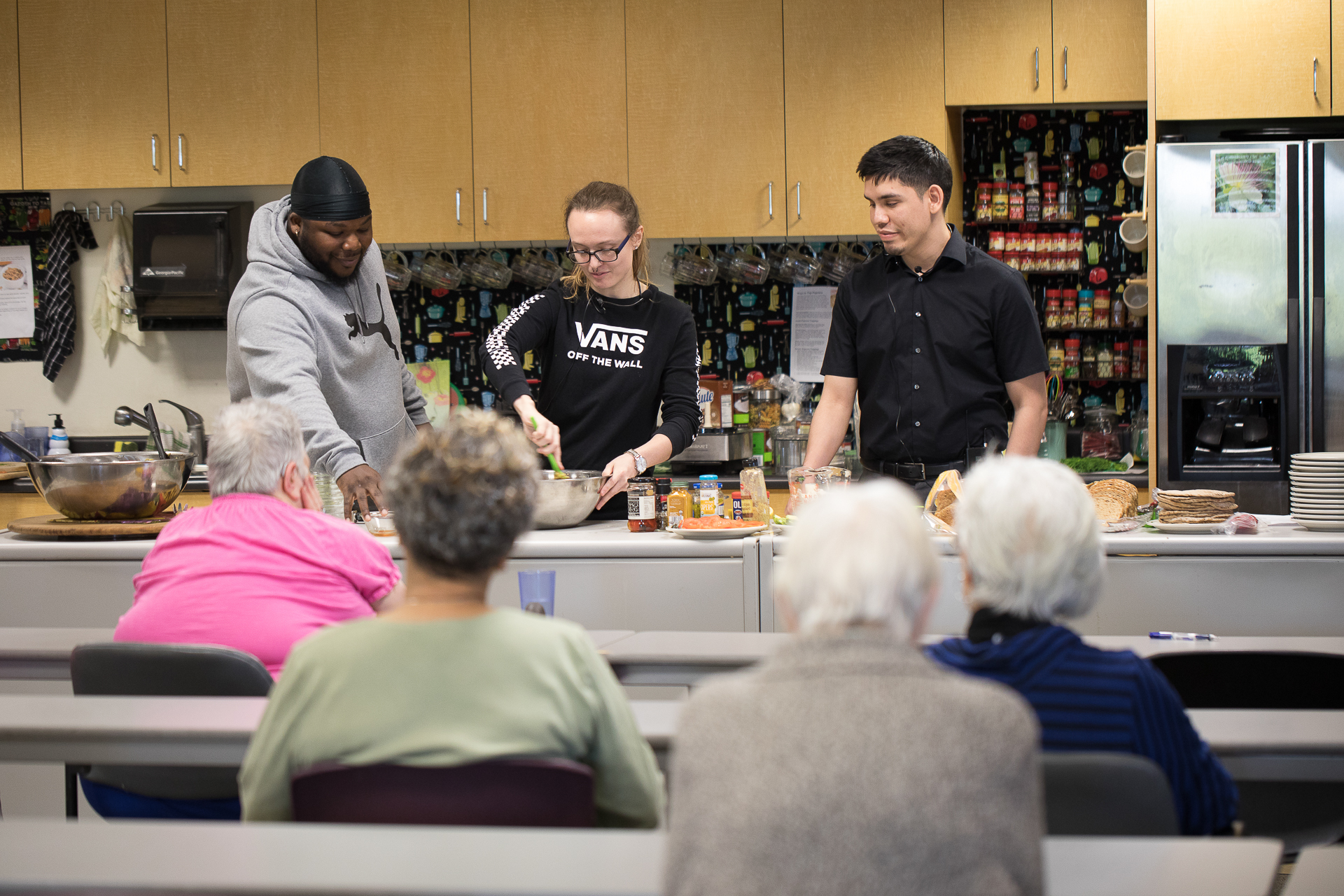 A Sac State student at a kitchen counter, stirring ingredients in a bowl, with two other students standing next to her, in front of an audience of seniors