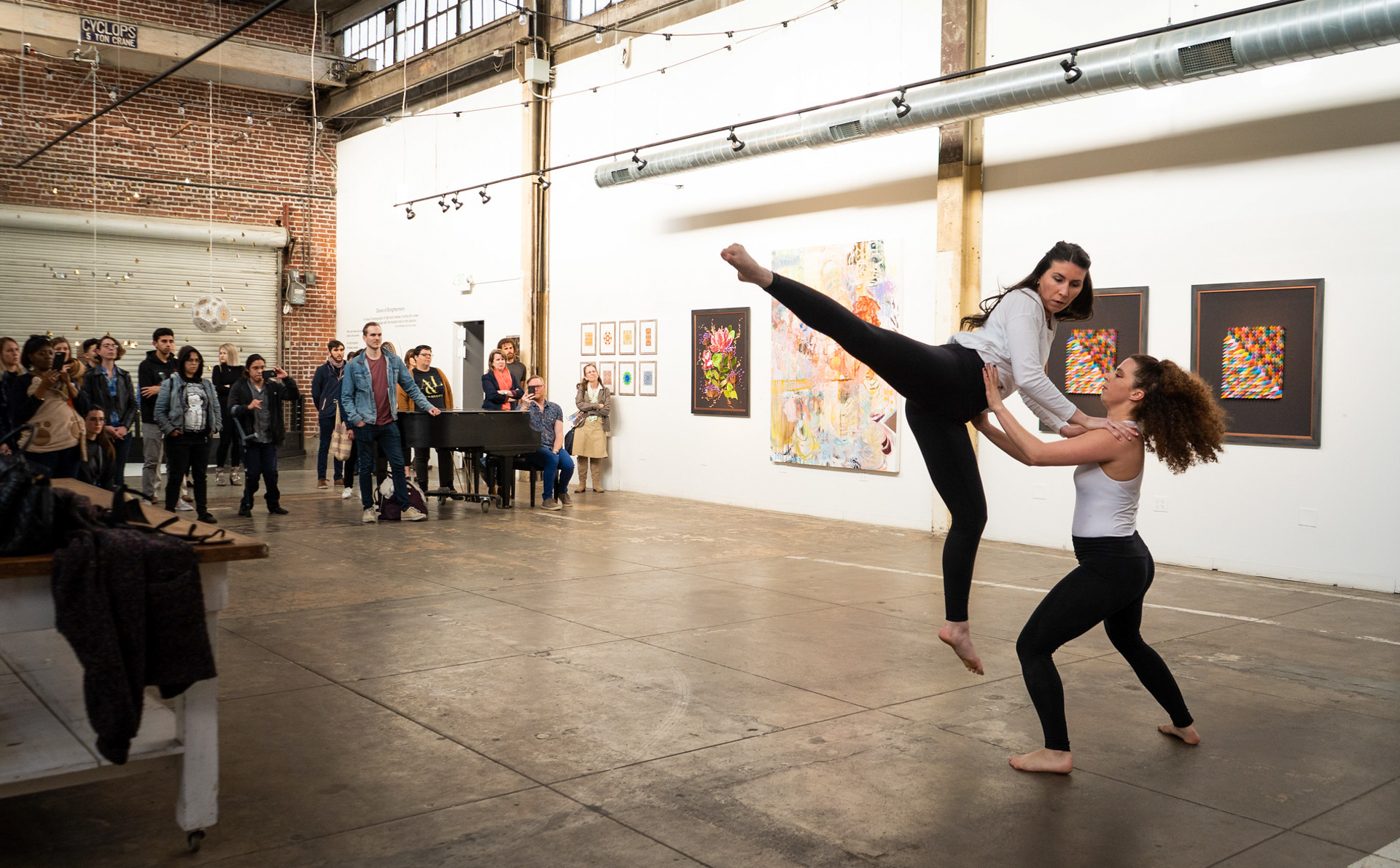 One dancer lifts another as audience members watch, indoors