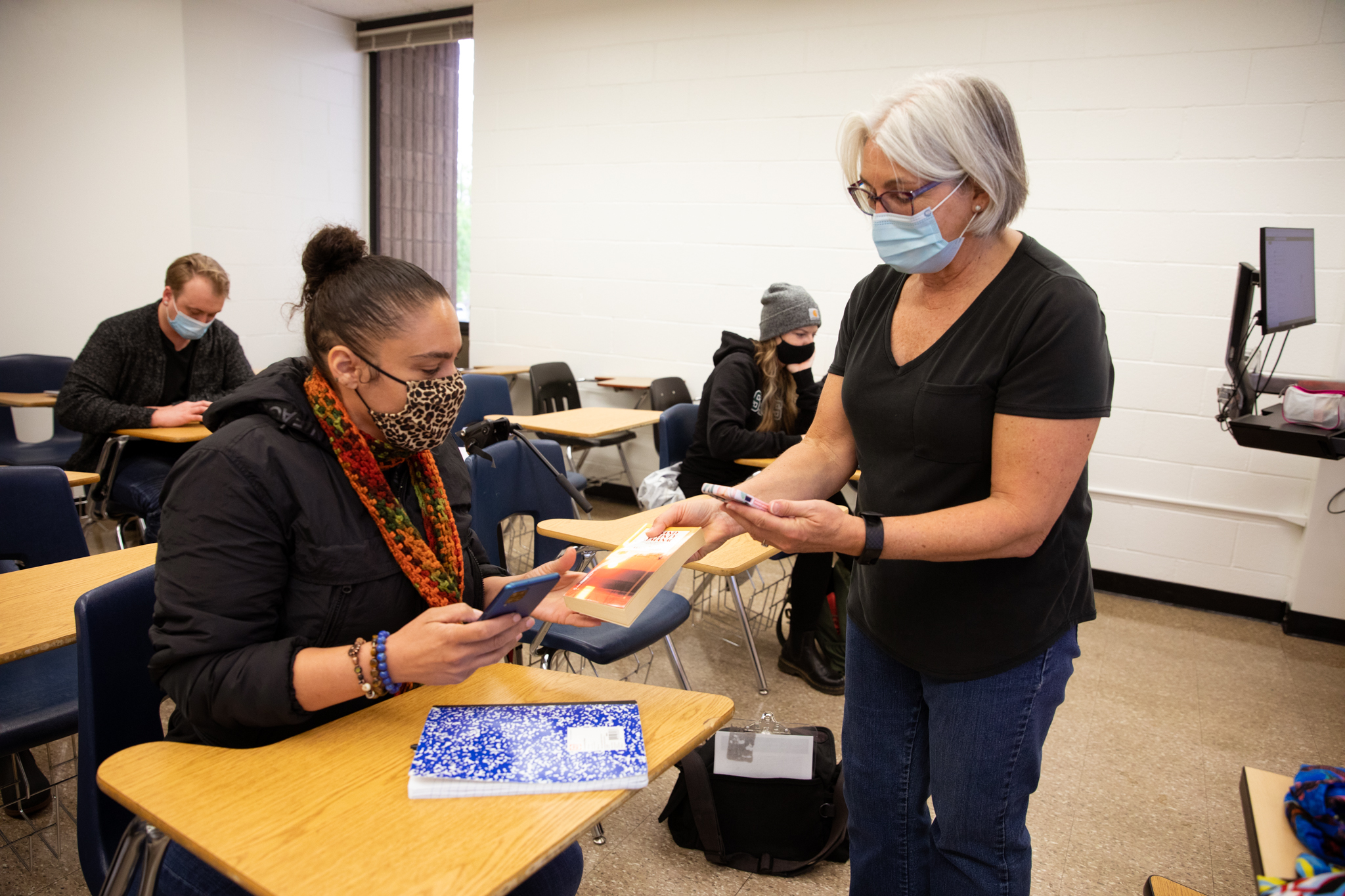 Two women, indoors in a classroom, holding smartphones, exchanging a book