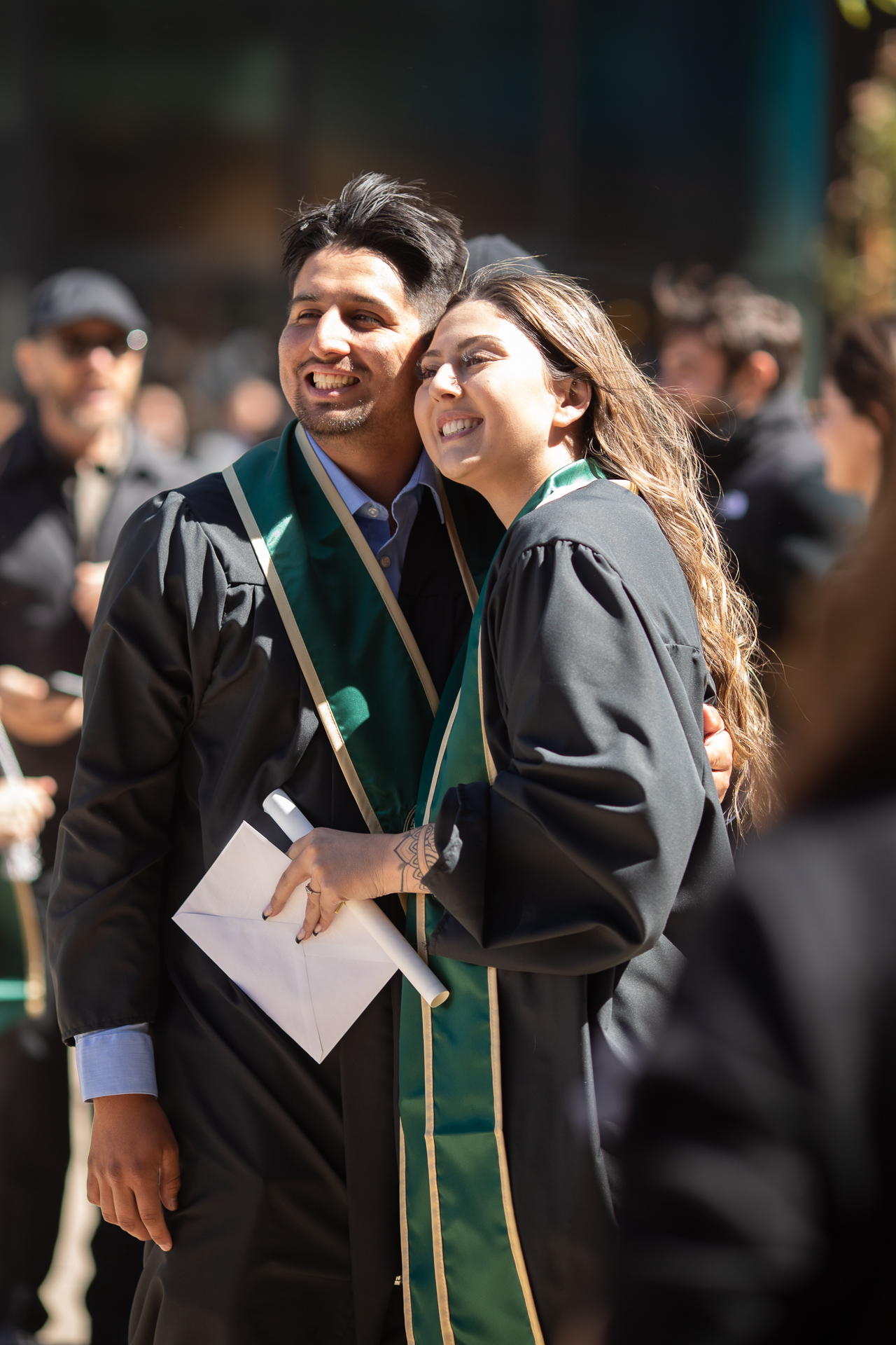 Two graduating students, outside, smiling, in academic regalia, one holding a diploma