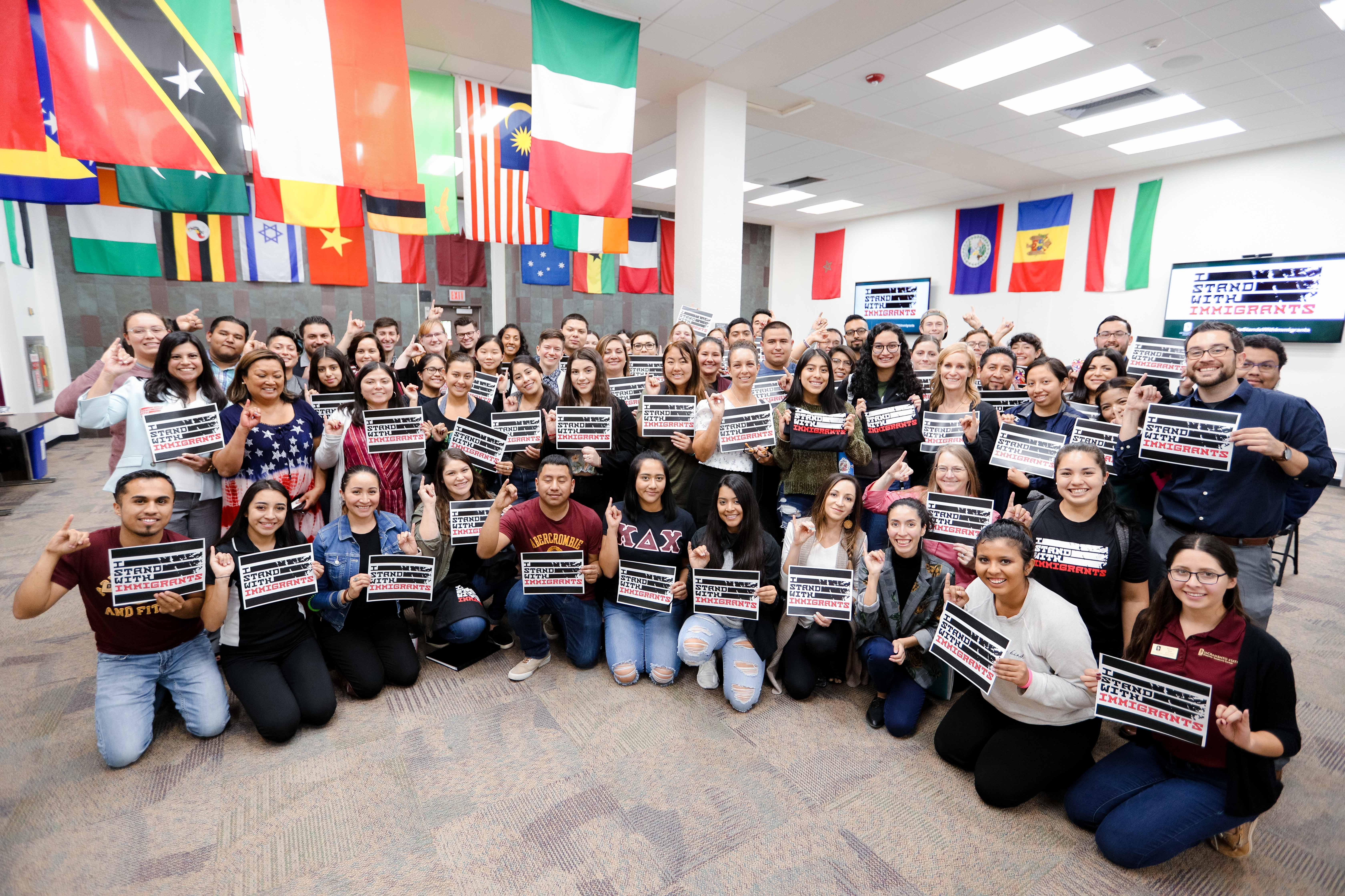 A large group of people, indoors, holding signs that read "I Stand With Immigrants"