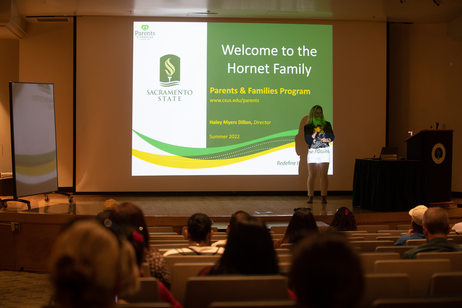 Haley Myers Dillon, standing on a stage in front of an audience, while a screen displays a slide reading "Welcome to the Hornet Family."