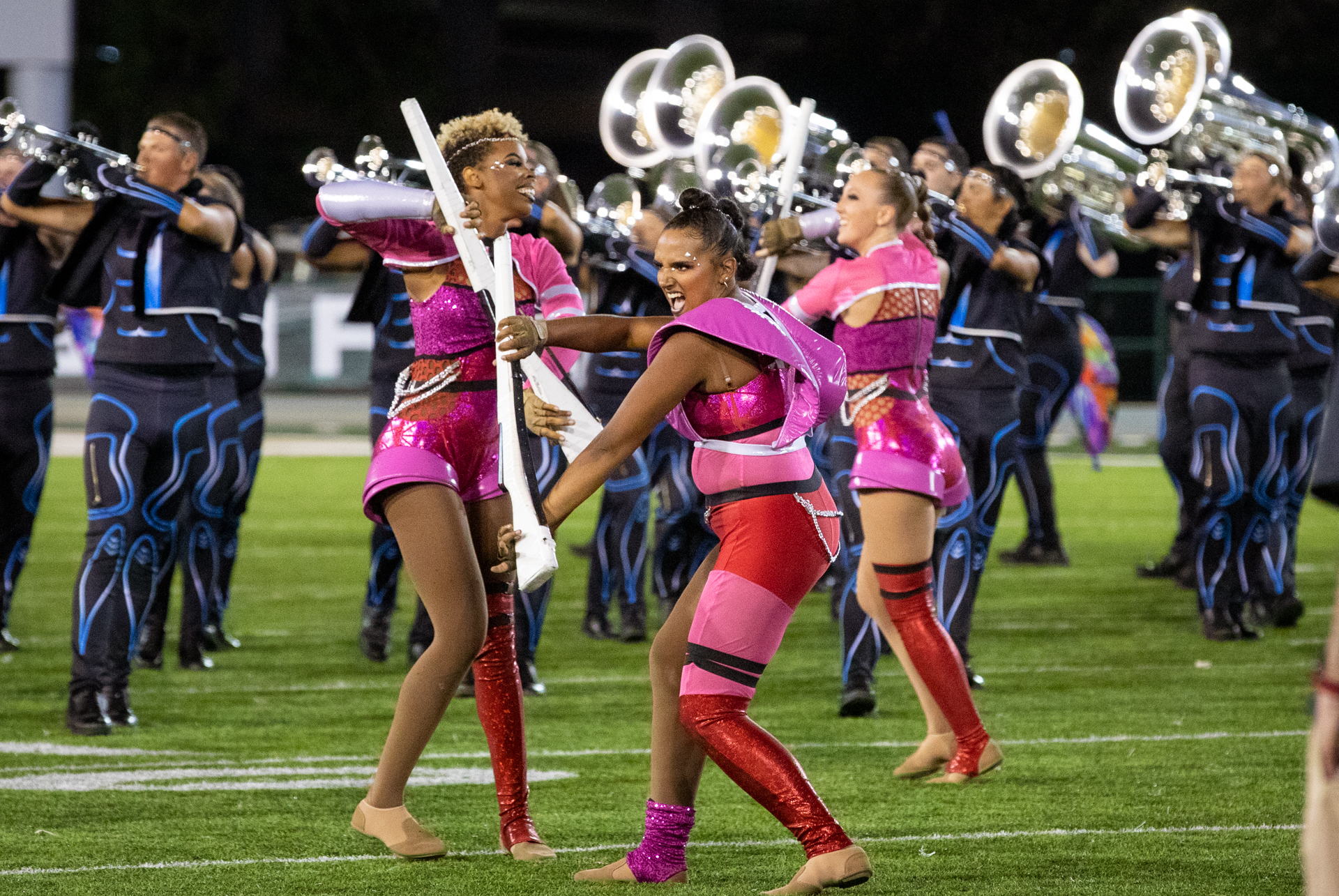 Members of the Mandarins drum and bugle corps dressed in lavish pink and blue costumes dance play instruments on the field at Sac State's Hornet Stadium.