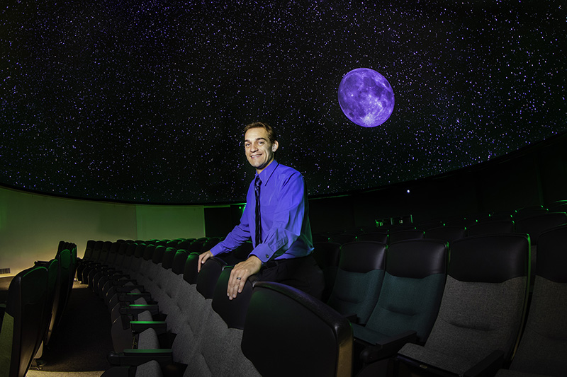 Physics Professor and Planetarium Director Kyle Watters stands against a backdrop of the earth and stars being broadcast on the dome inside the Planetarium.