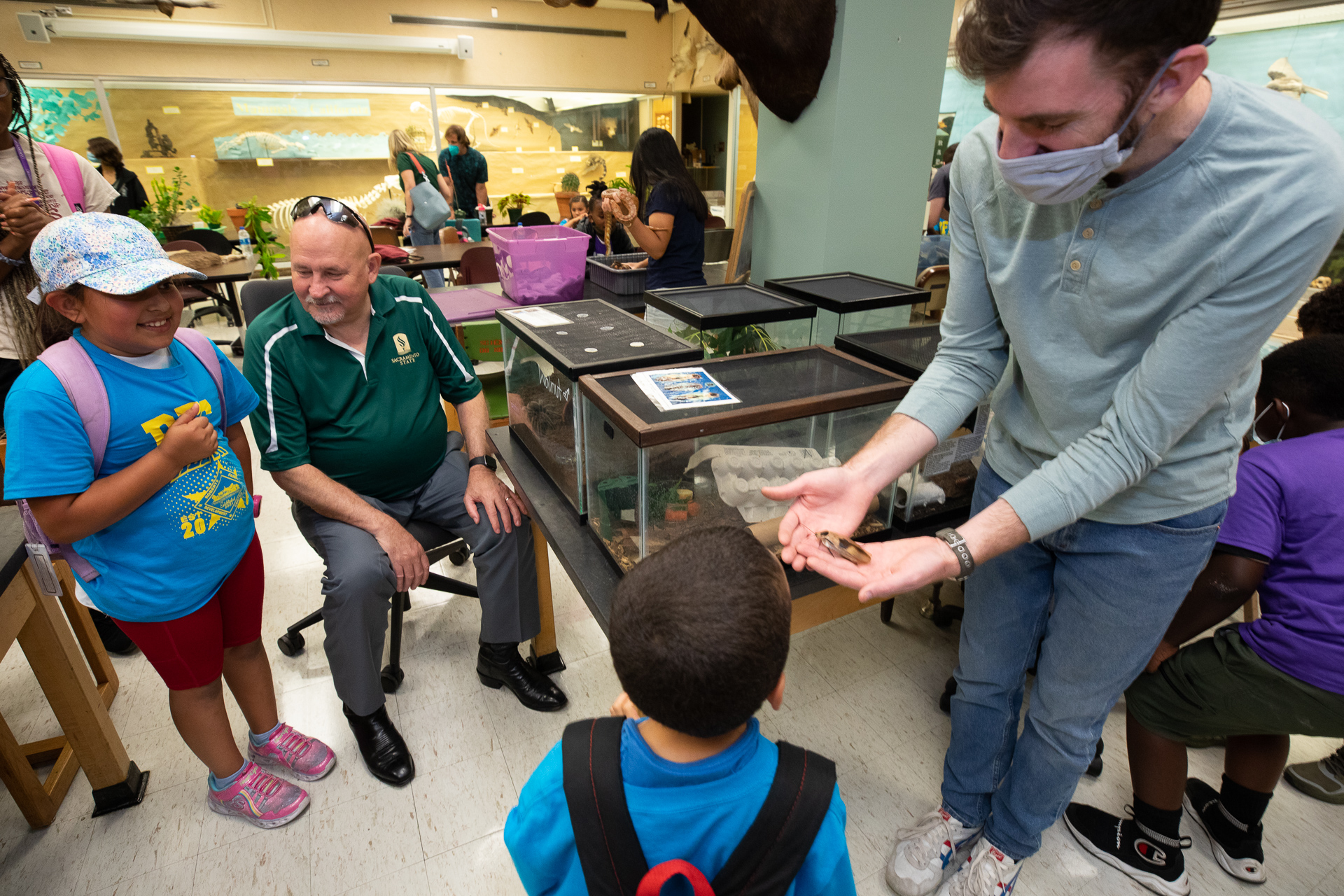 President Robert S. Nelsen, wearing a green Sac State shirt, sits with Roberts Family Development Center students as they observe a large brown insect at the Vertebrate Museum in Sequoia Hall.
