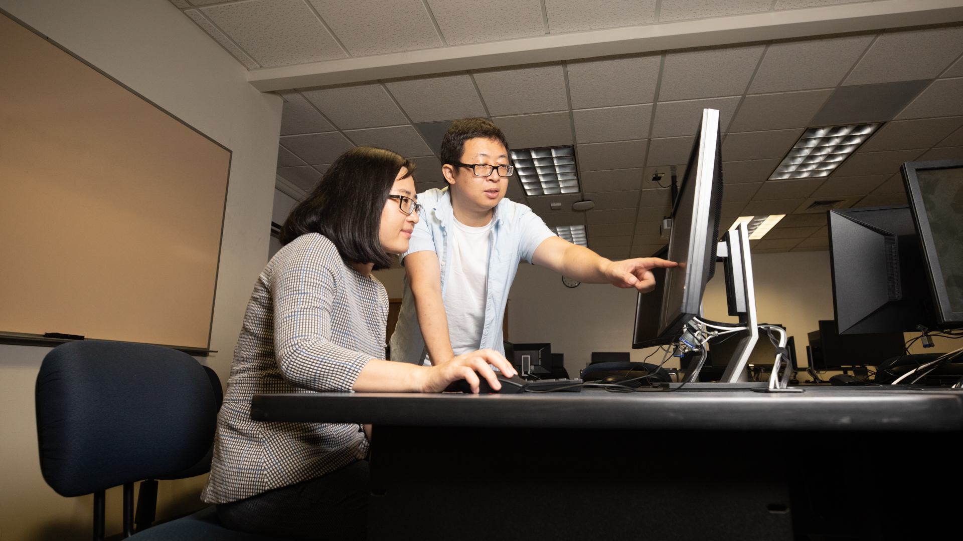 Sacramento State Computer Science professors Xiaoyan “Sherry” Sun (sitting down) and Jun Dai work together on a project in a campus computer lab as Sun controls the mouse while Dai points at the screen.