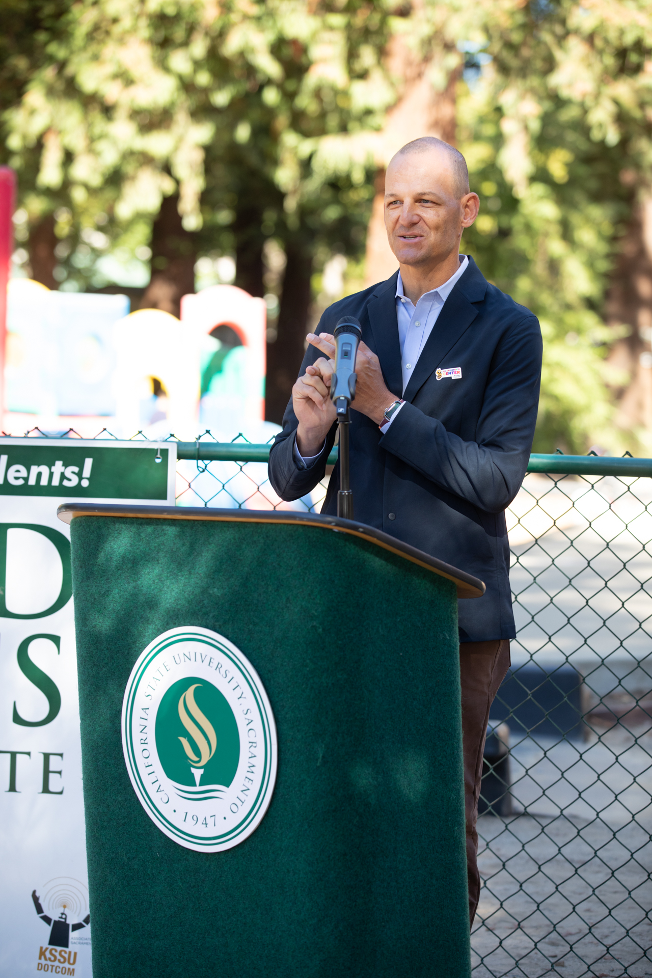 California Assemblymember Kevin McCarty speaks behind a podium in front of the Sacramento State ASI Children's Center playground.