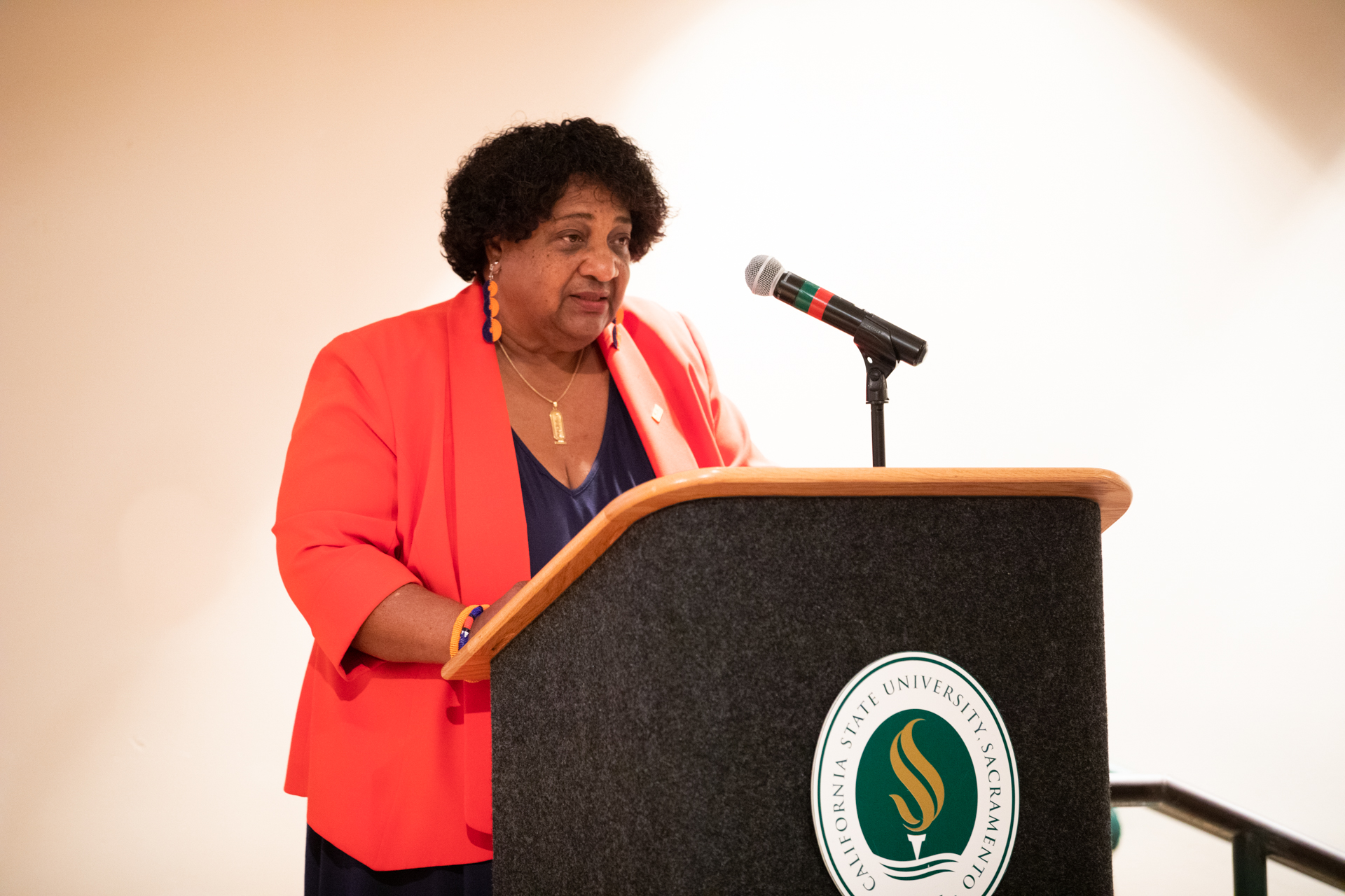 California Secretary of State Shirley N. Weber, wearing a vibrant red jacket, speaks at a podium during a recent voter rights forum at Sac State's Hinde Auditorium.