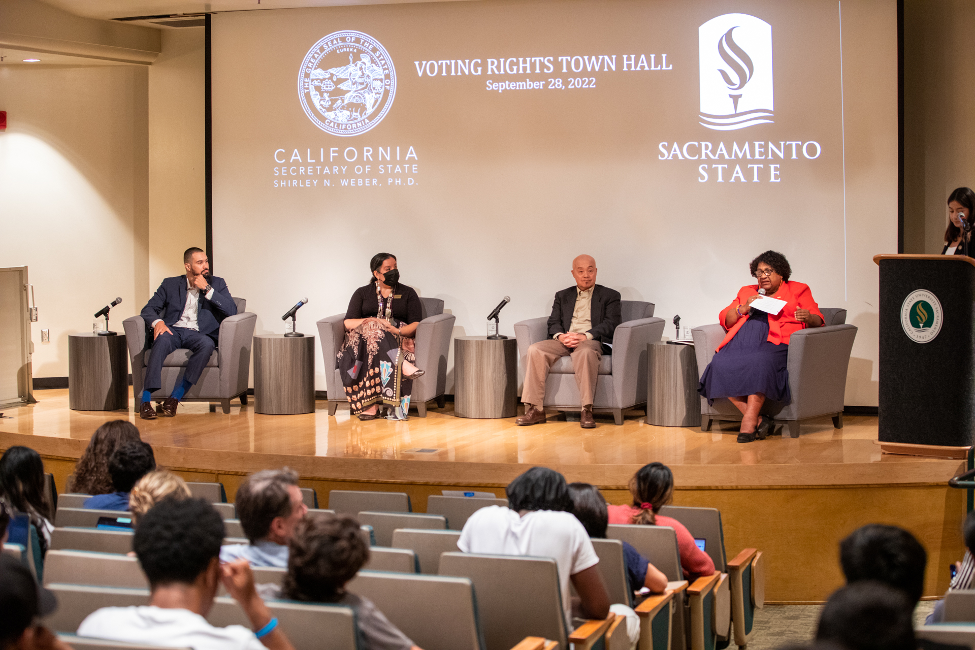 Sacramento State faculty members and California Secretary of State Shirley N. Weber, red jacket, sit in chairs on stage at Hinde Auditorium while ASI President Salma Pacheco speaks at the podium at a voter rights forum. 