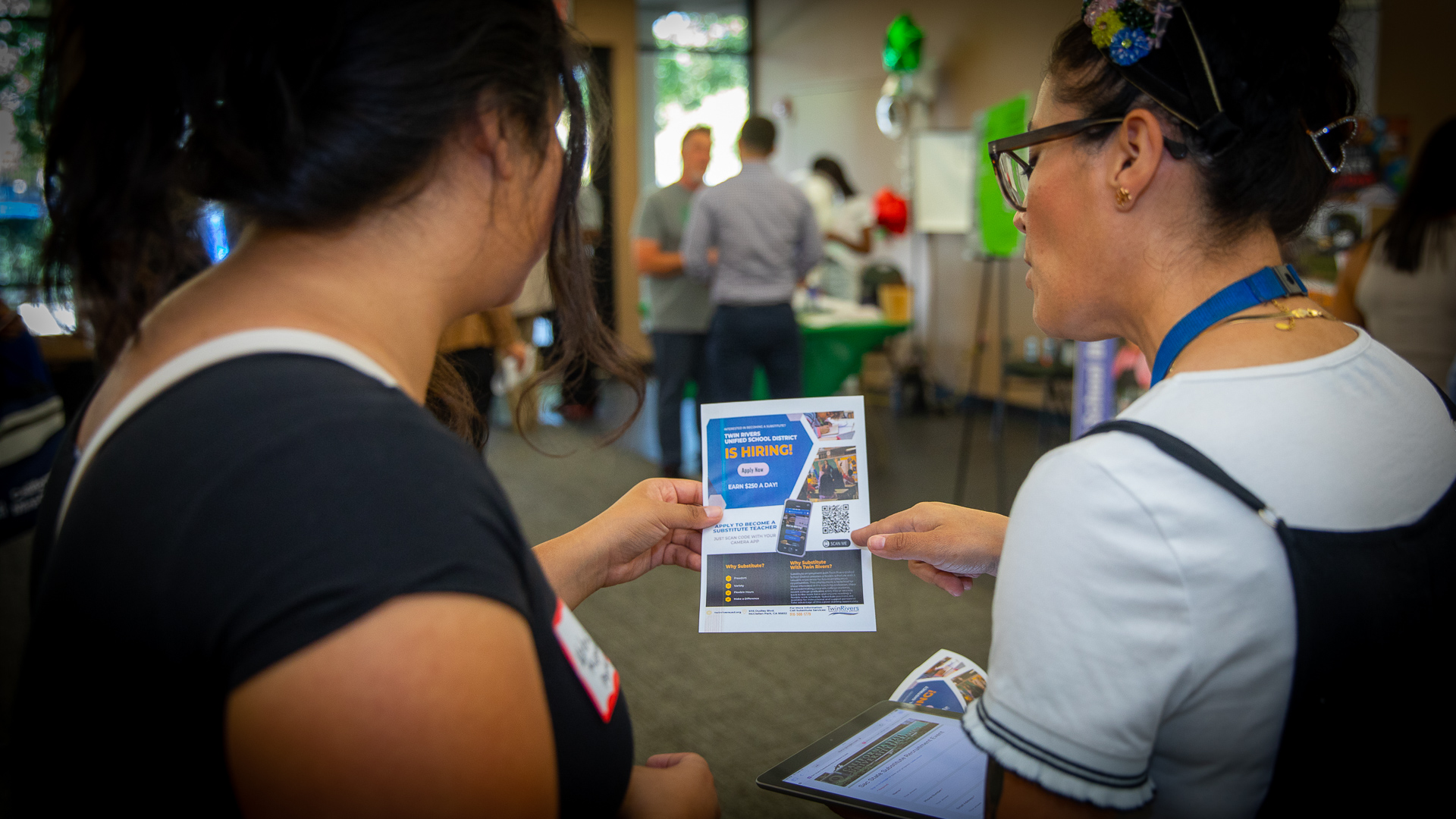 Recent Sac State graduate Valeria Miranda, wearing a black shirt, speaks to a representative of Twin Rivers Unified School District, who is wearing a light blue top and overalls, while the pair hold a substitute teacher hiring information sheet.