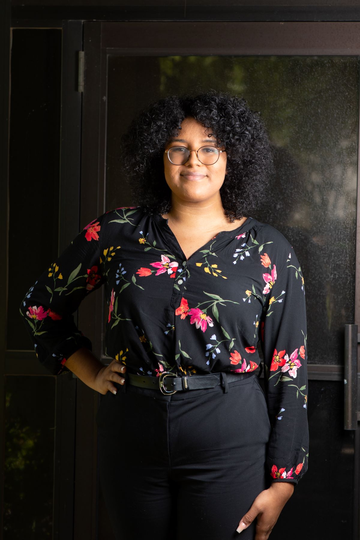 Kenya Burton, 2022 CSU Trustees’ Award for Outstanding Achievement recipient, poses with one hand on her hip on a black studio background wearing a black shirt adorned with flowers.