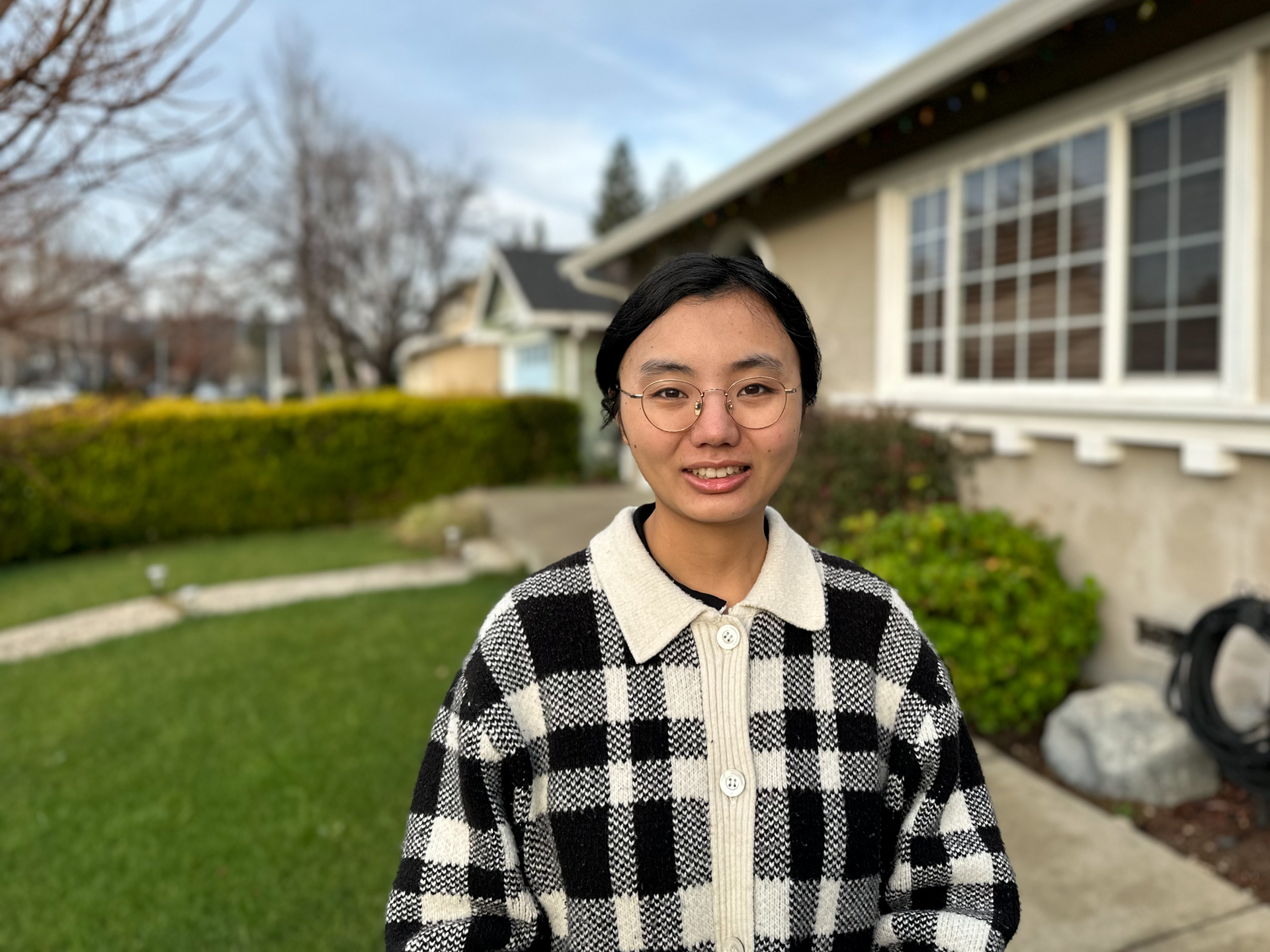 Assistant Professor of Communication Studies Cheng Hong poses on the backdrop of green grass and a light-colored house. 