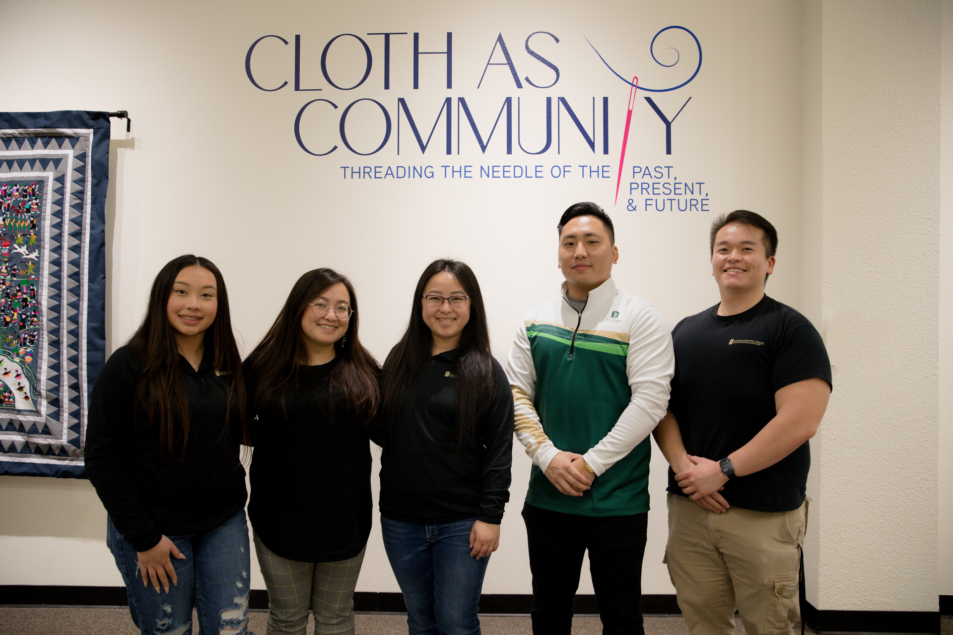 A group shot, standing under a "Cloth as Community: Threading the Needle of the Past, Present and Future" sign, of some of the people who worked on a new Hmong clothing exhibit at Sac State.