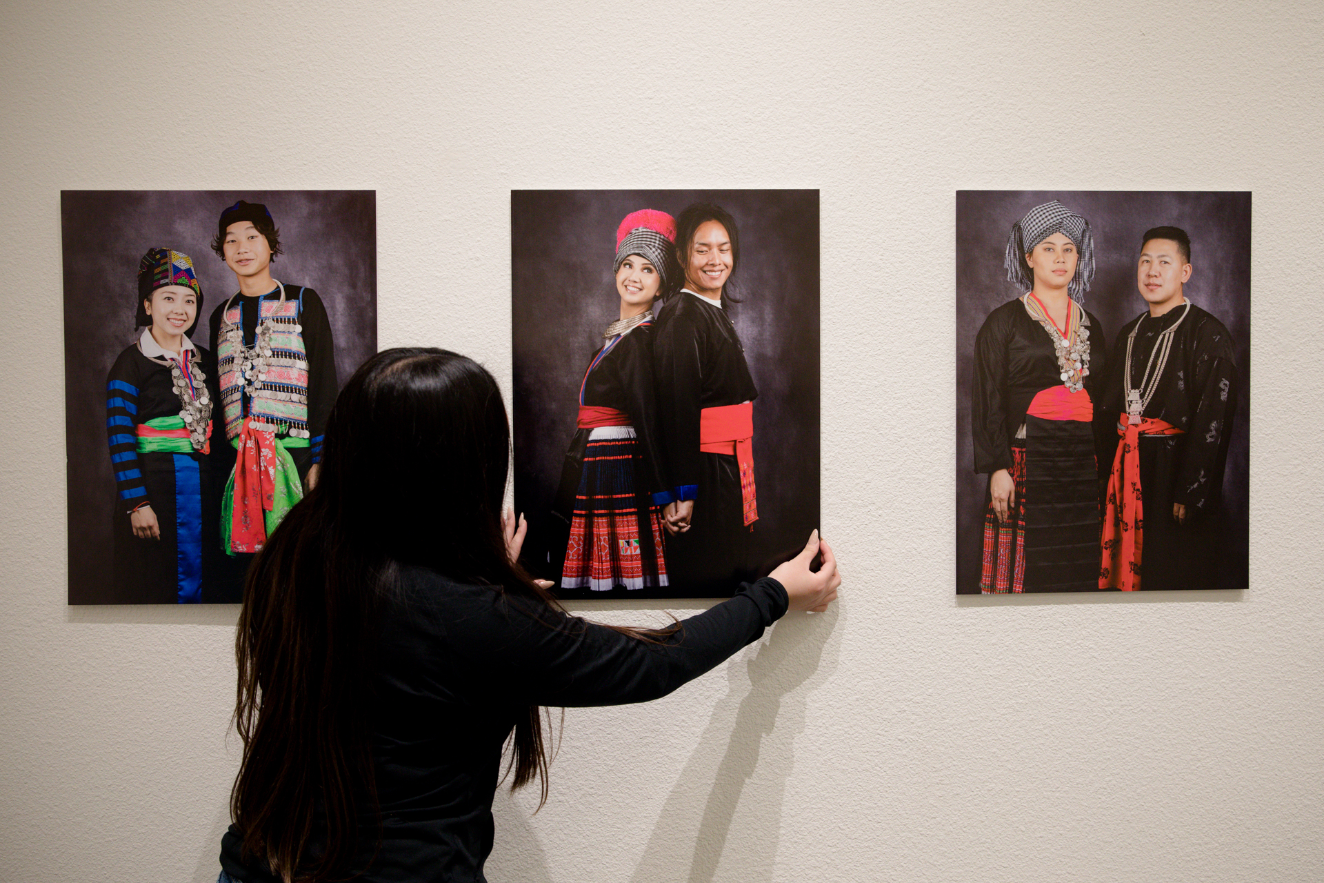 A woman adjusts prints on a wall of men and women modeling traditional Hmong attires, on display as part of an exhibit at Sac State.