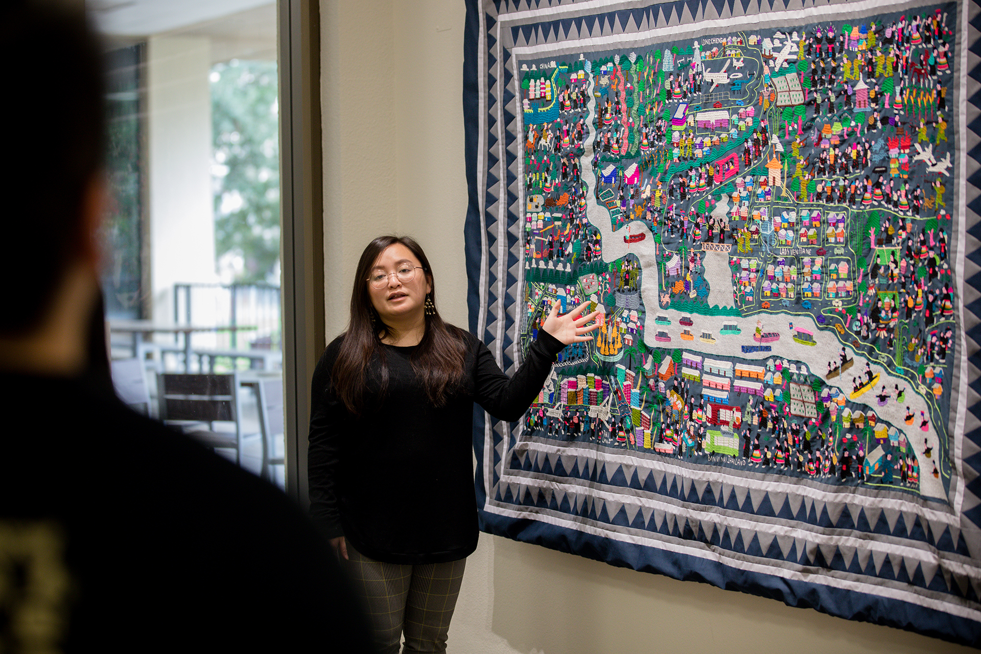 A woman tells a spectator about a large traditional Hmong story cloth, which hangs on the wall.