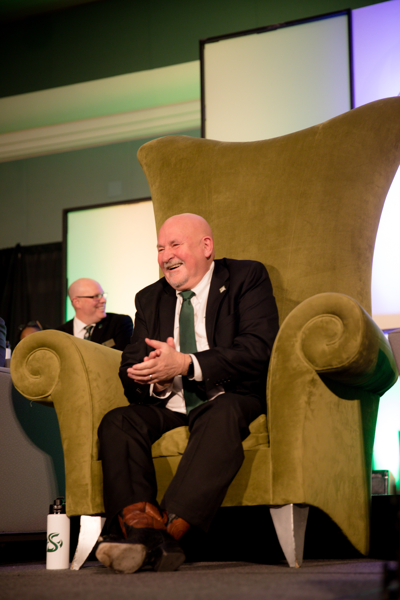 Sitting in an oversized green chair, President Nelsen laughs and claps during a joke at his roast.