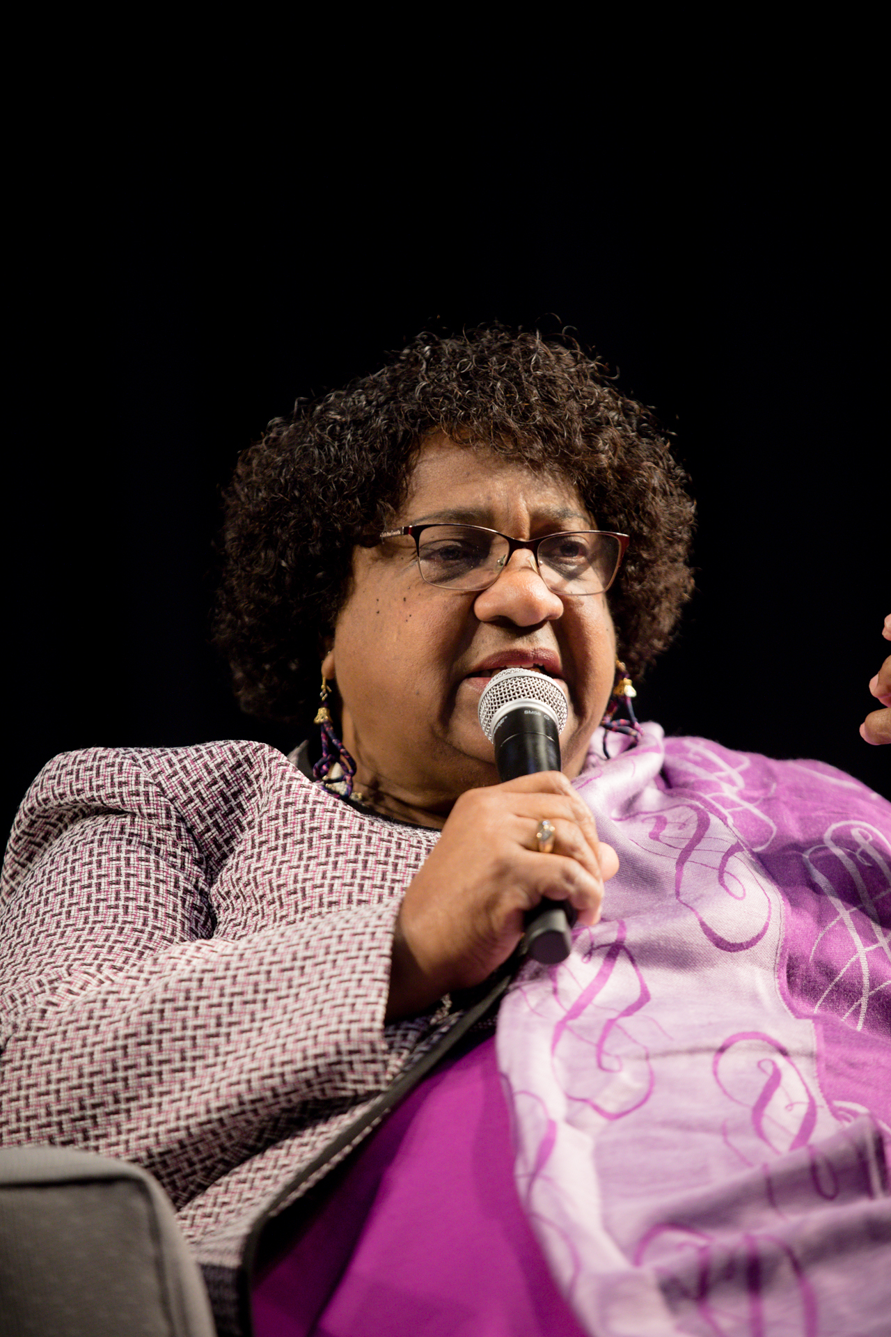 California Secretary of State Shirley Weber wearing a purple outfit and holding a microphone during a speech.