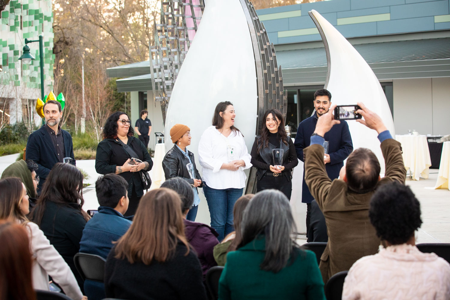 Several students and alumni hold awards in front of Tributary, a public art display. 