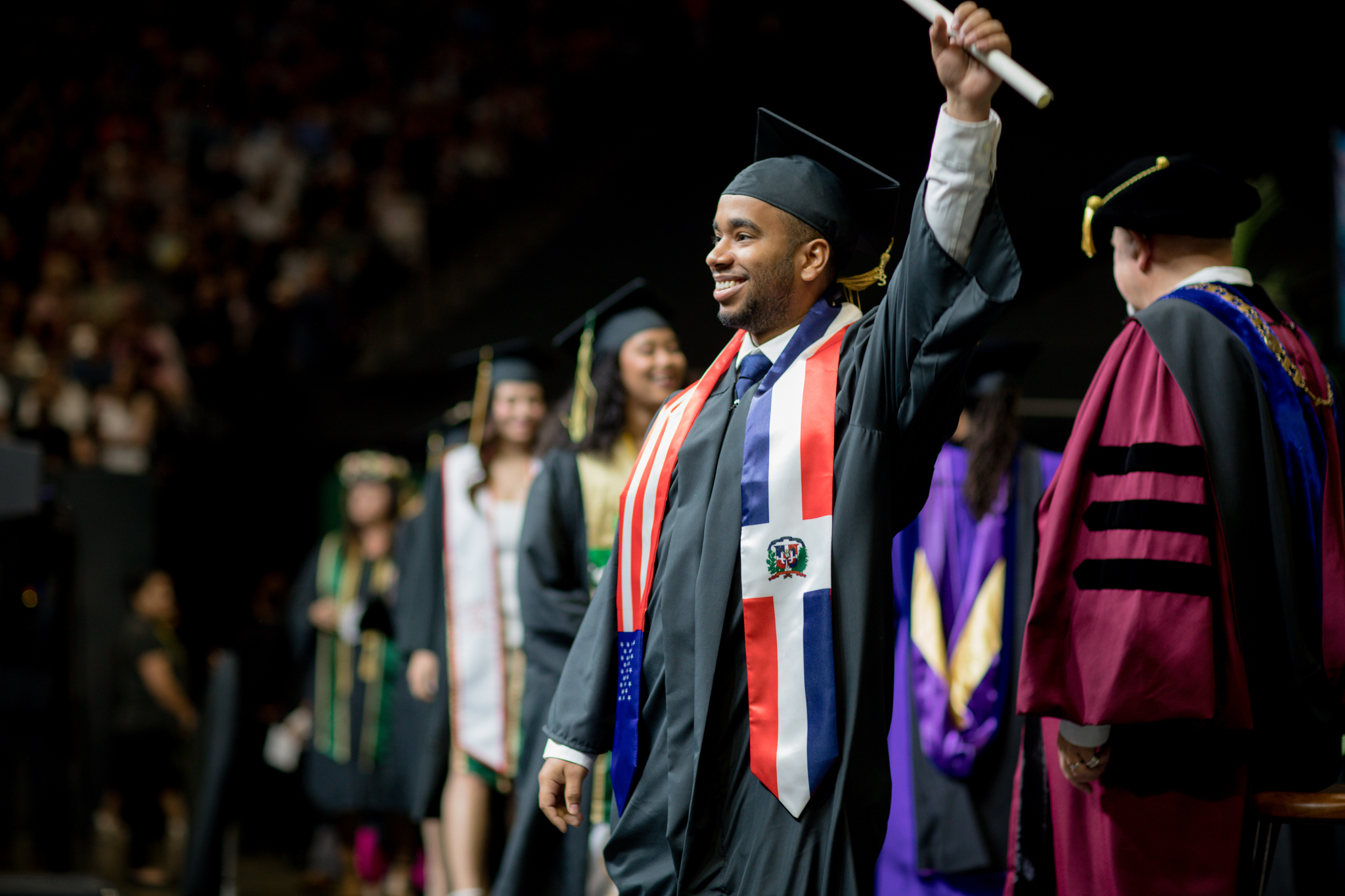 A male Sac State student, in graduation regalia, stands on the Commencement stage looking at the audience