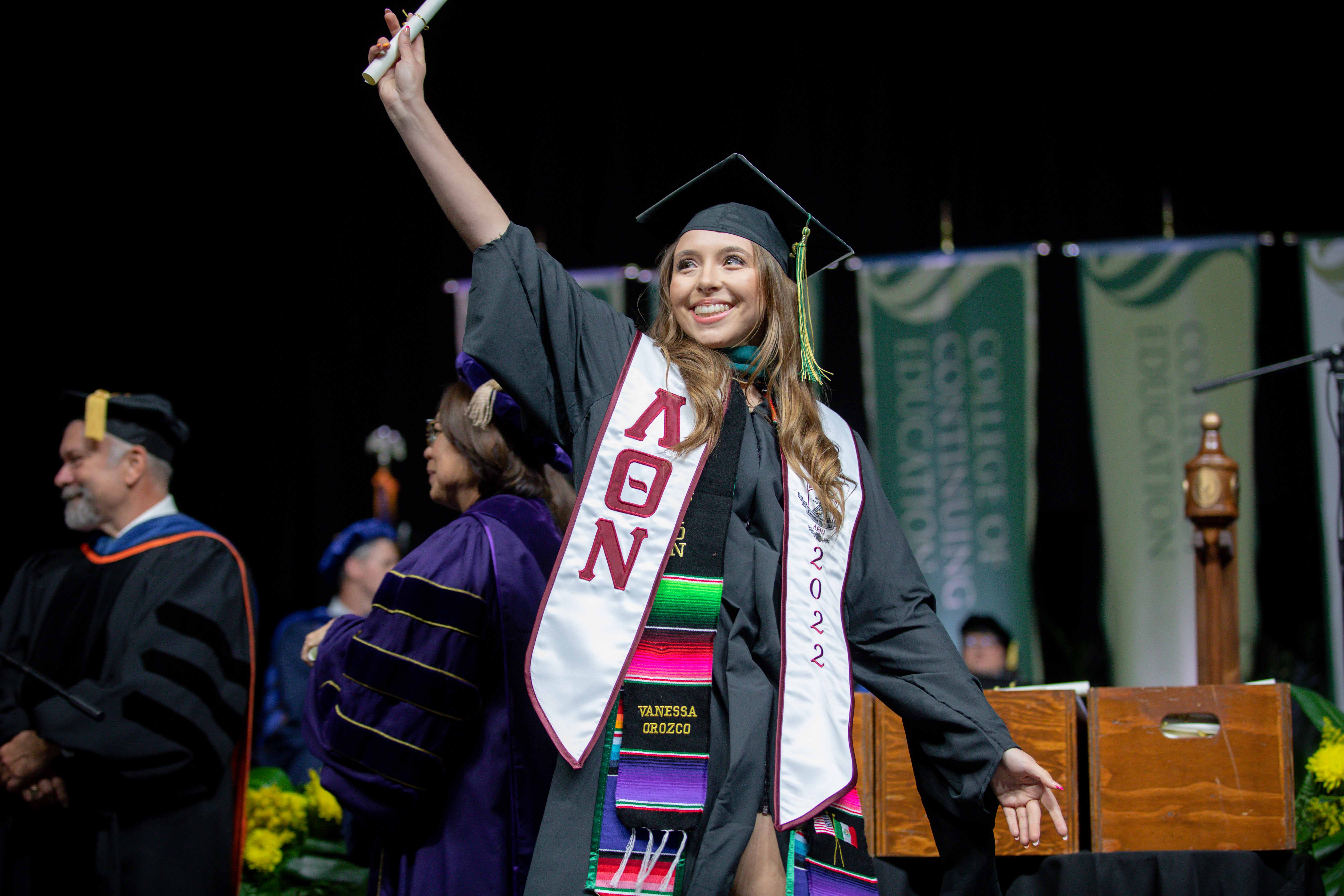 A woman in academic regalia, standing on the Commencement stage, smiling and holding her diploma aloft